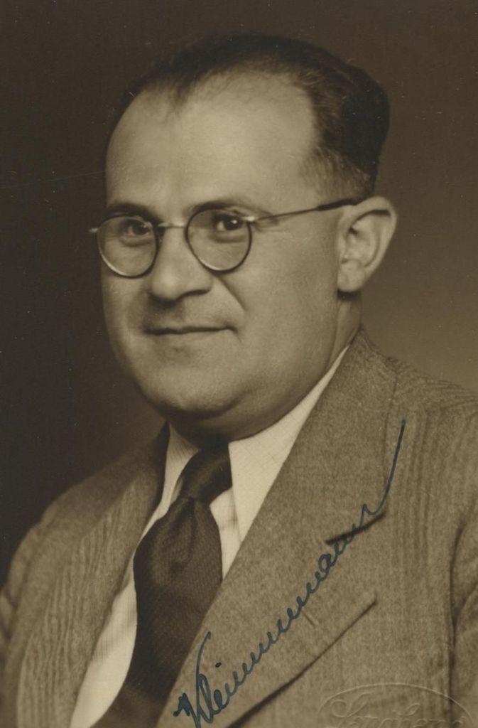 Portrait of a man, featuring a close-up of a smiling individual wearing glasses, a suit, and tie.