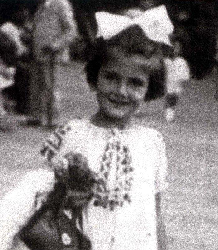 Young girl smiling at the camera, wearing a light-colored dress with decorative trim and a large bow in her hair.