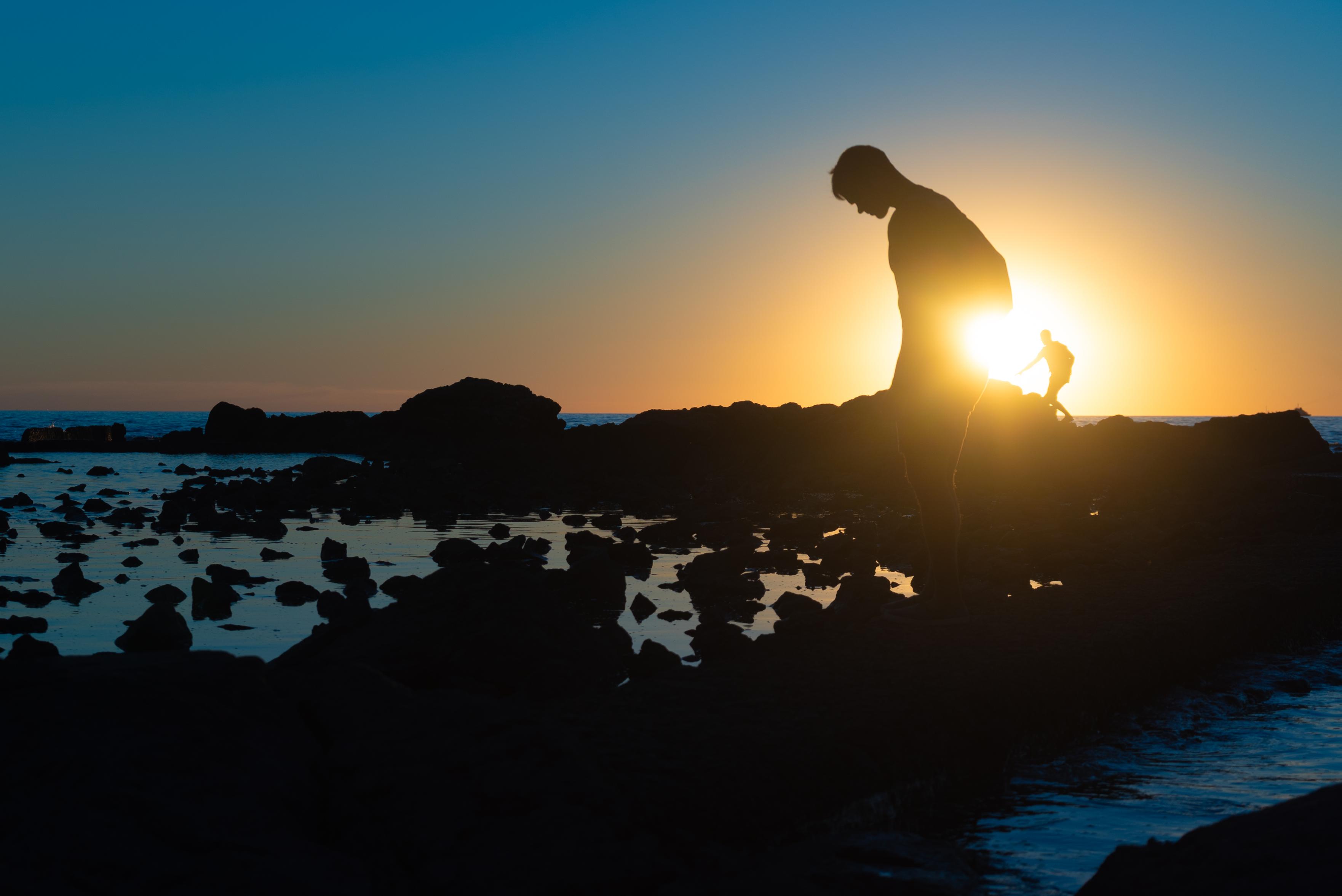 black silhouette of a rocky shore and two men against a sunset sky. on eof the men is closer to the foreground, navigating the rocks and another is in the back. the sun is just between them, blowing out some of the image in warm yellow tones and the rest of the sky transitions into a beautiful blue towards the edges