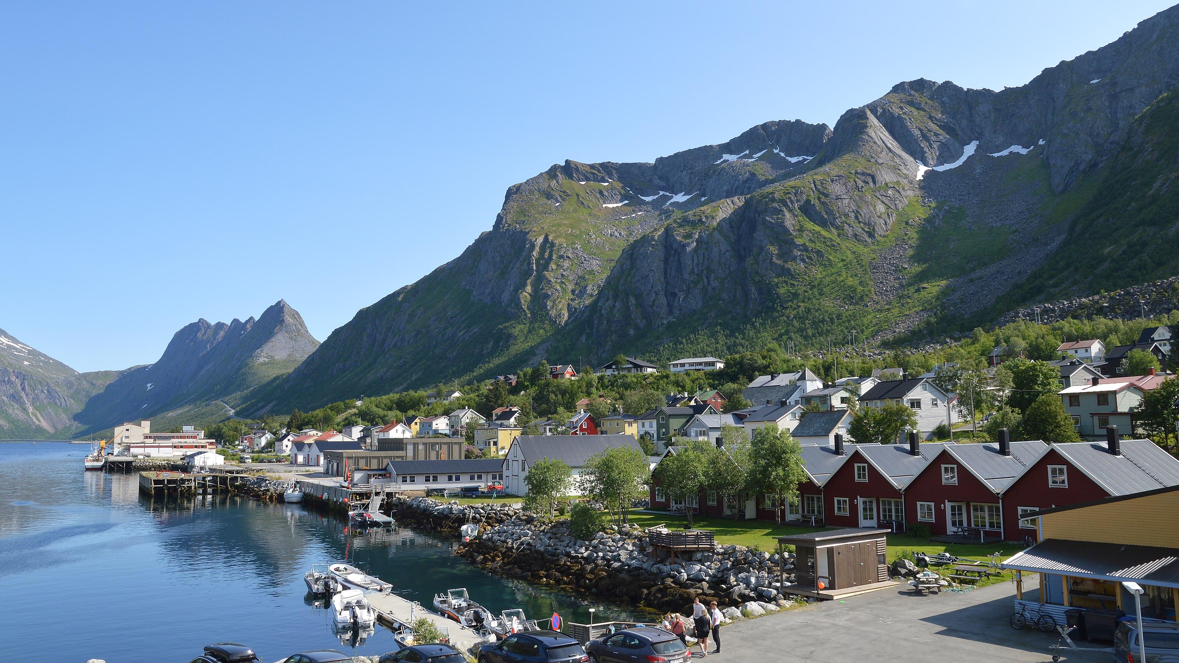 A photo of a small town on the water with mountains behind it. The sky is clear and blue.