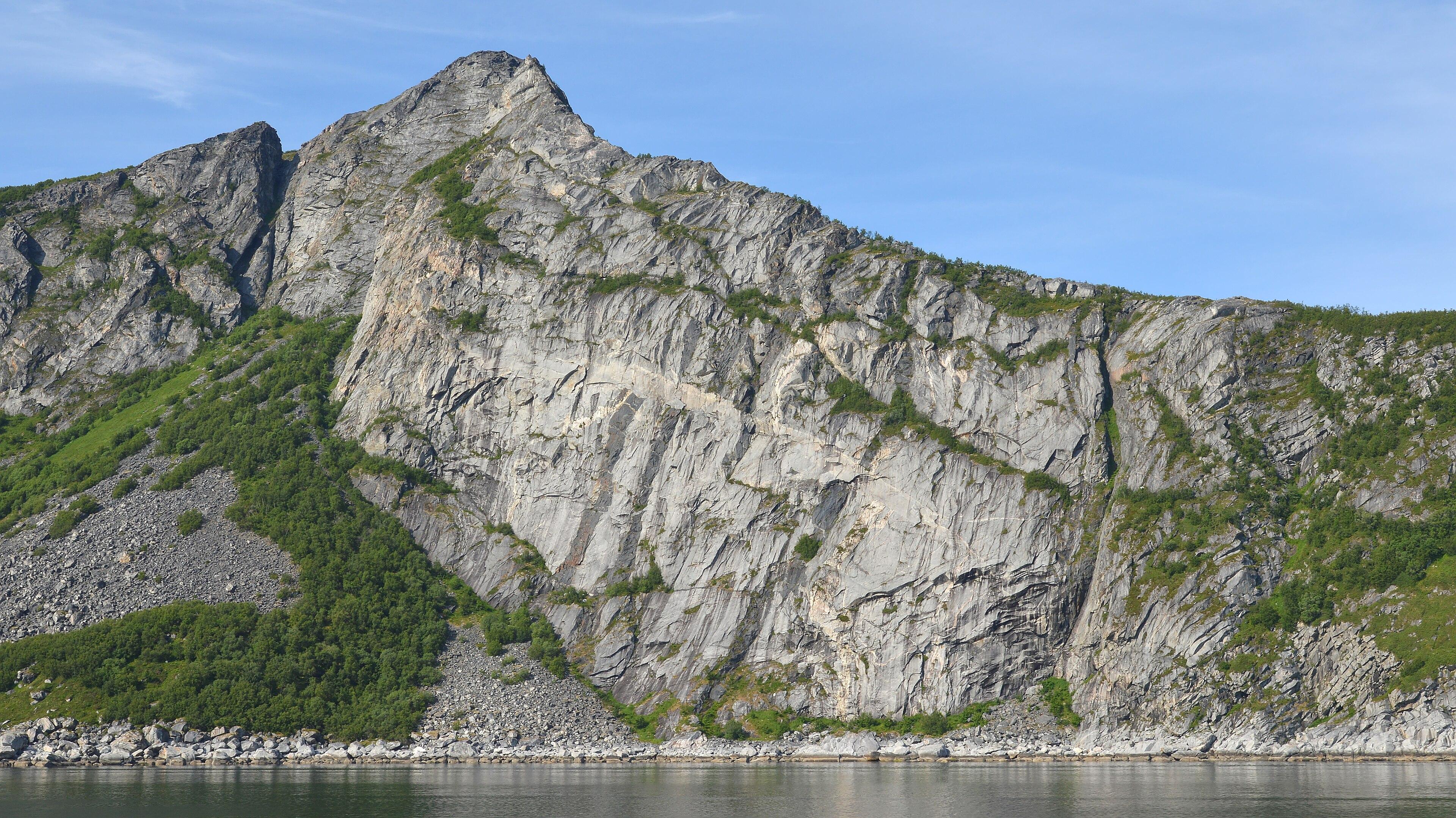 A photo of a mountain leading into water. The sky is blue and mostly clear.