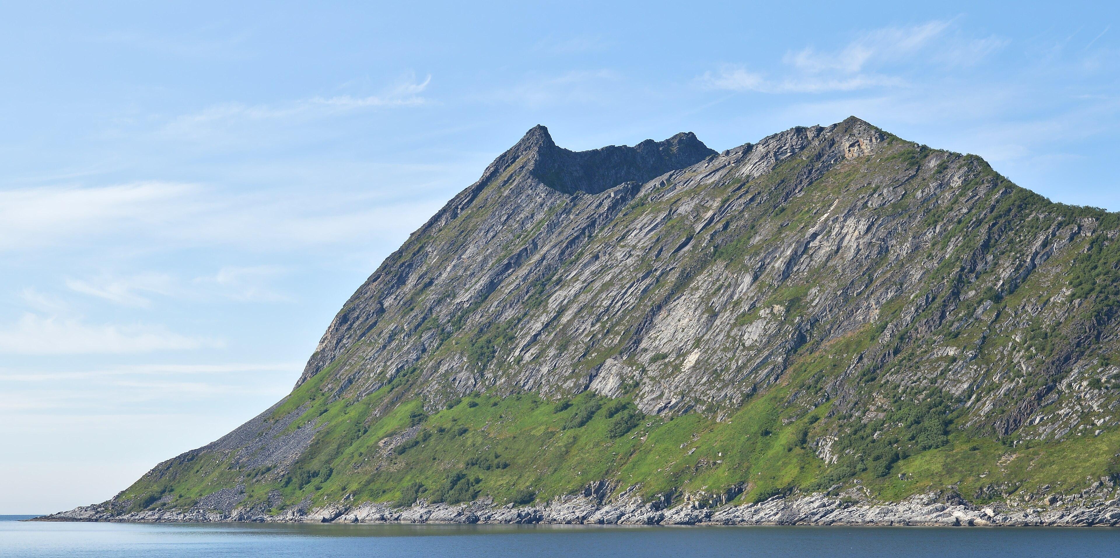 A photo of a mountain leading into water. The sky is blue and with wispy clouds in it.