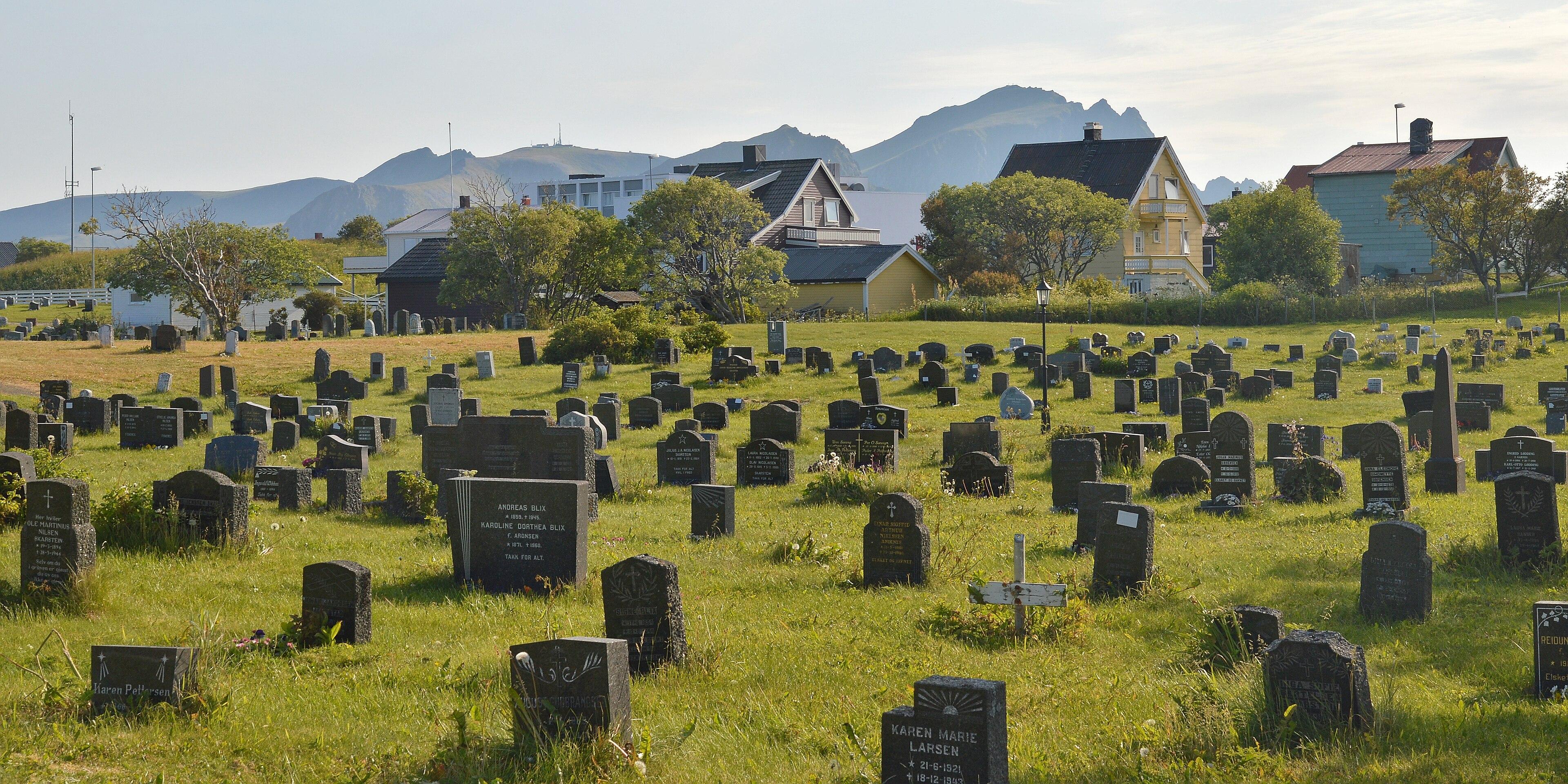 A photo of a graveyard with houses behind it and mountains beyond that. The sky is a hazy blue with some faint clouds.