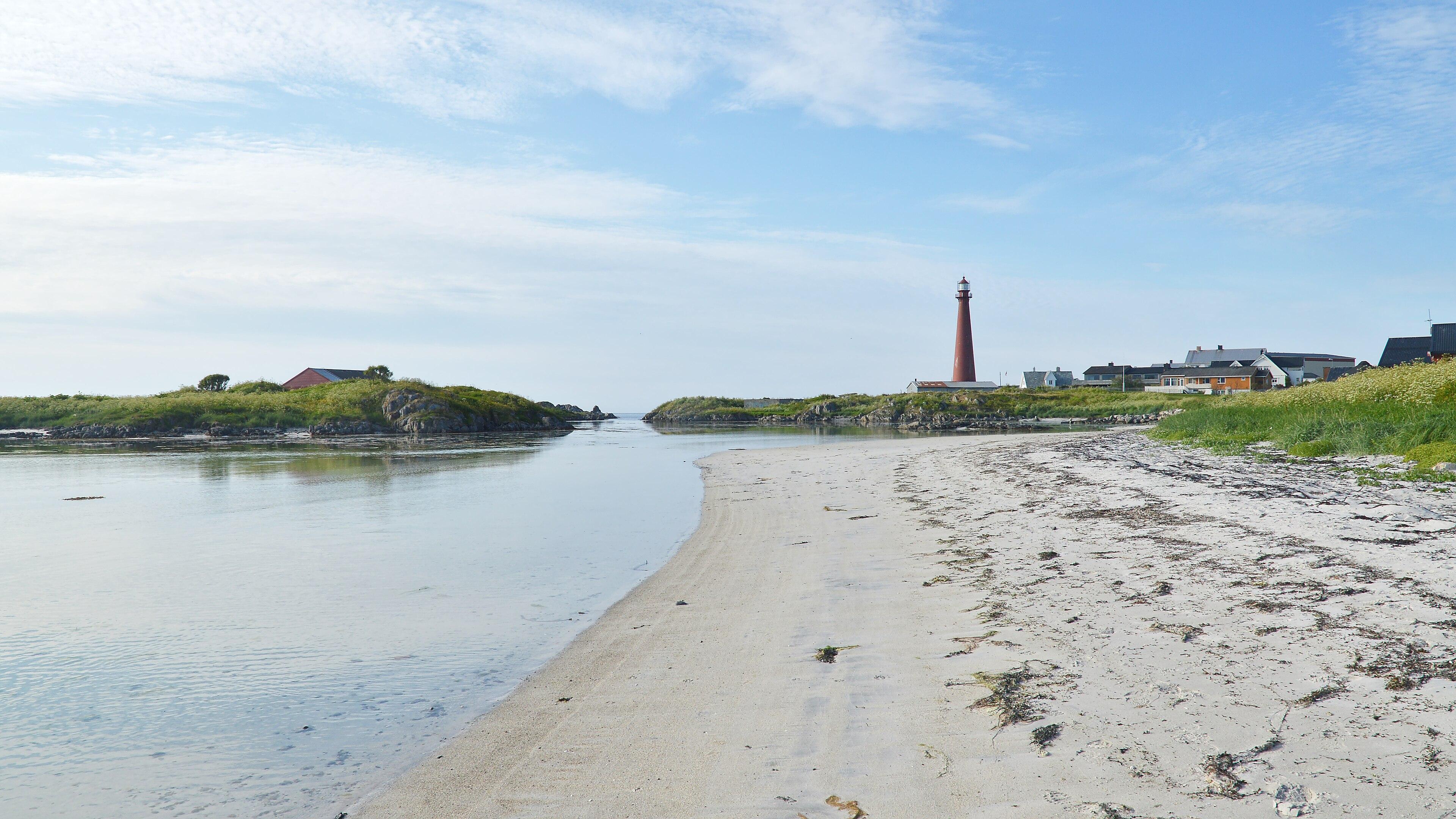 A photo of a sandy beach wit ha lighthouse in the distance. There are several small islands in the water. The sky is blue with white clouds in it.