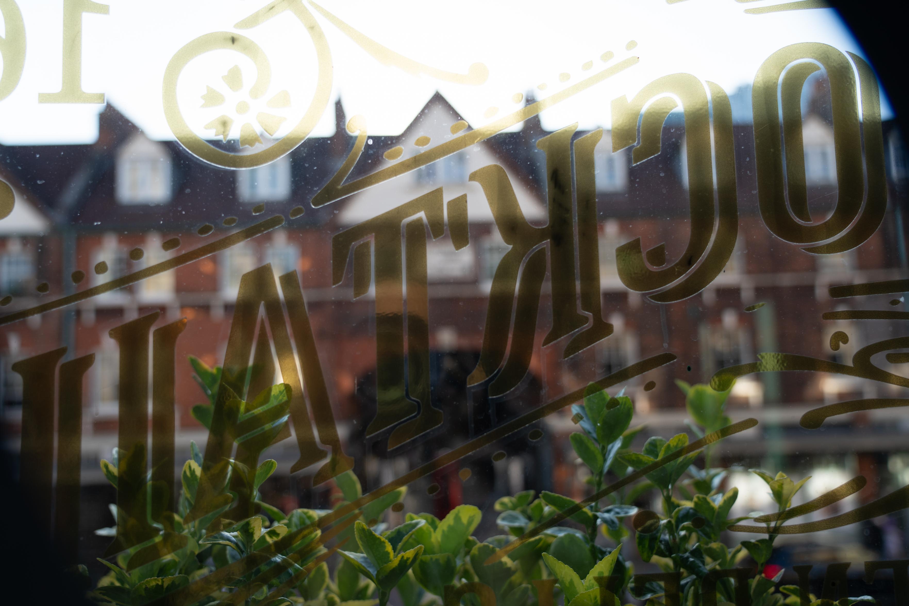a photo, looking out through a pub window in London. gold Victorian letters read "COCKTAIL" in reverse. through the window, the blurred red brick facades of a typical British street. a small plant on the windowsill.