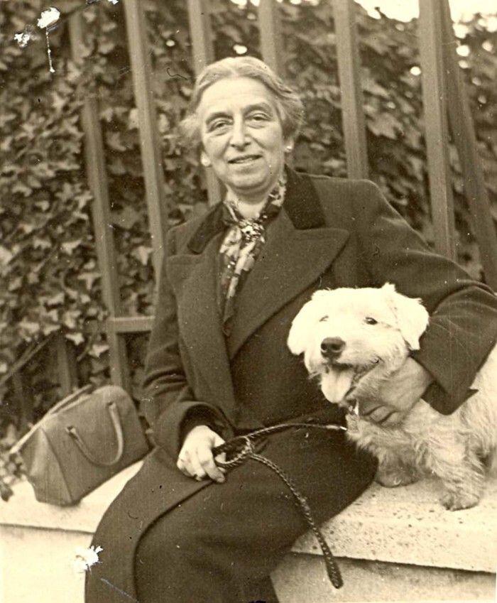 A vintage photograph of a woman sitting on a low wall, smiling slightly and looking directly at the camera. The woman is dressed in a dark coat and a patterned scarf, with a small handbag beside them. They are affectionately holding a small, light-colored dog with a leash