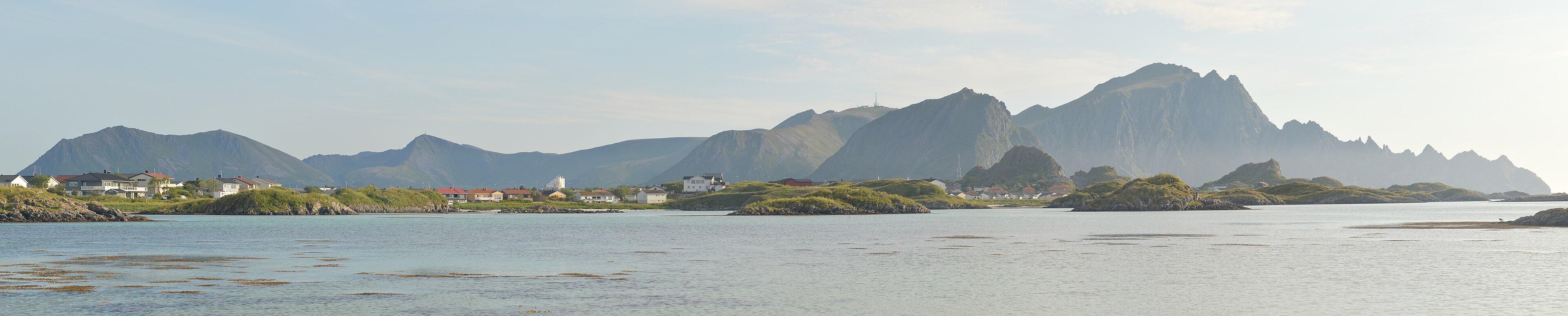A panoramic photo of jagged mountains behind low green islands. There are houses along the shore. The sky is hazy and with clouds in it.