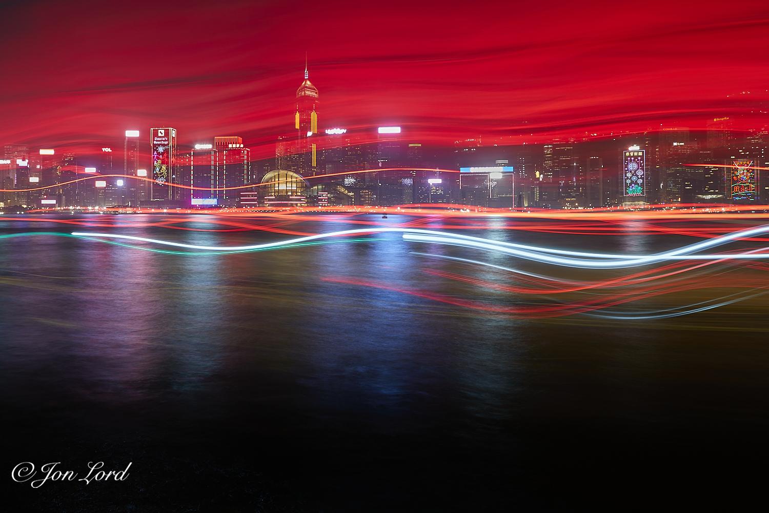 This is a colour nighttime cityscape, long exposure, photo of a body of water with the light trails of passing boats and a high-rise waterfront cityscape in the background. Victoria Harbour, Hong Kong (2018).

The dark harbour water rises from the base of the photo upwards to a little above the half way up level. The water at the base is pitch black with it becoming an undulating silver and multi-coloured tone from the reflections of the bright lights of the distant, about one to two km away, high-rise waterfront cityscape. From the quarter up level to the top of the photo are a series of random wavy  predominantly red lines crossing the image horizontally from left to right: the light trails caused by passing boats and a long camera exposure. Around the centre section of the photo, the lines are much brighter and clearly defined, with the red coloured lines towards the top having a dimmer less distinct, almost mist like appearance. Beyond, is the city's waterfront lined with multiple, brightly lit skyscrapers, many with large and illuminated signs on their roofs. Some with lighting in the shape of Christmas decorations stretching the height of those buildings, one with 'Seasons Greetings' in large neon lettering near the top. The rapidly rising terrain behind the waterfront is obscured by the misty light trails towards the top of the image.