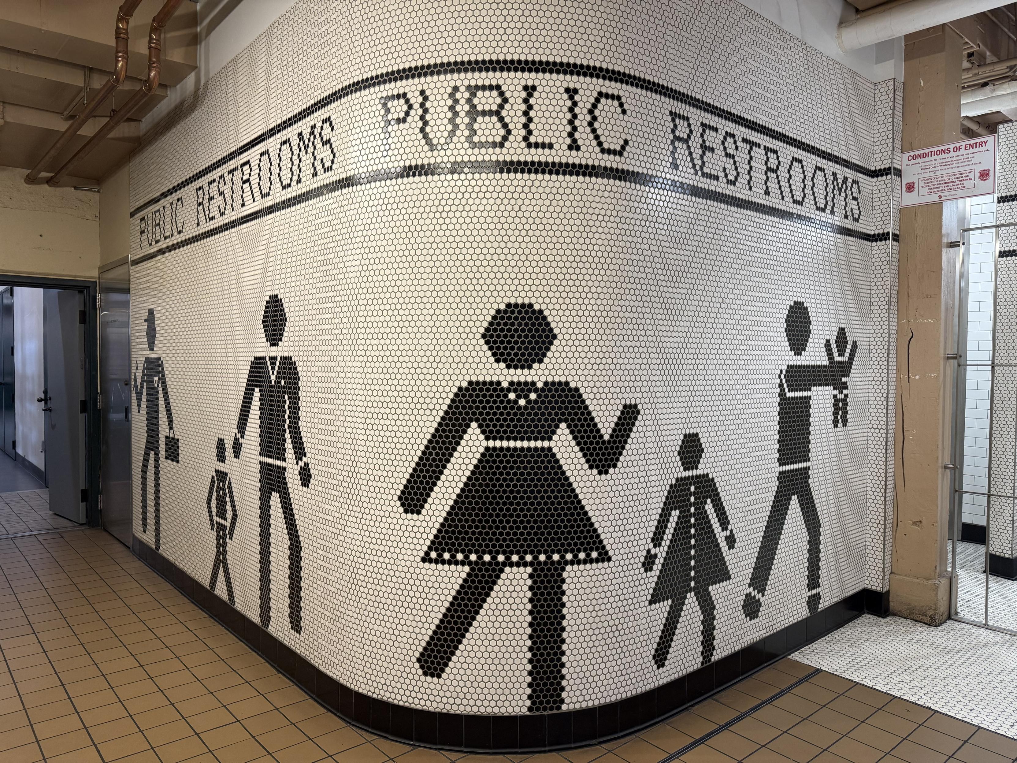 A tiled wall in a restroom area featuring black and white hexagonal tiles with silhouettes of people, including an adult man, an adult woman, and children. The words "PUBLIC RESTROOMS" are displayed above the graphics, indicating the location.