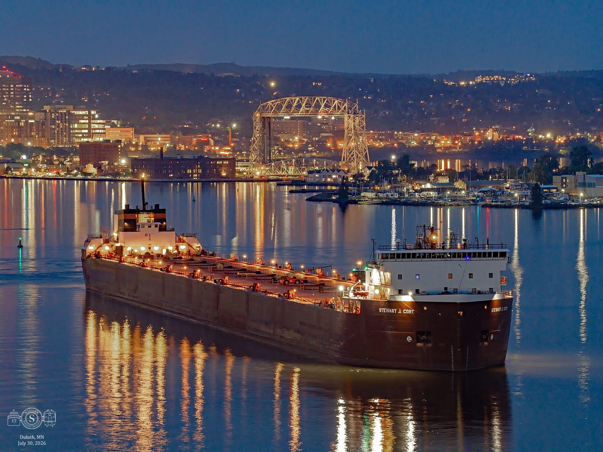A photo of a night time shot of a Great Lakes freighter waiting for fuel with some of the lights of Duluth skyline in the background
