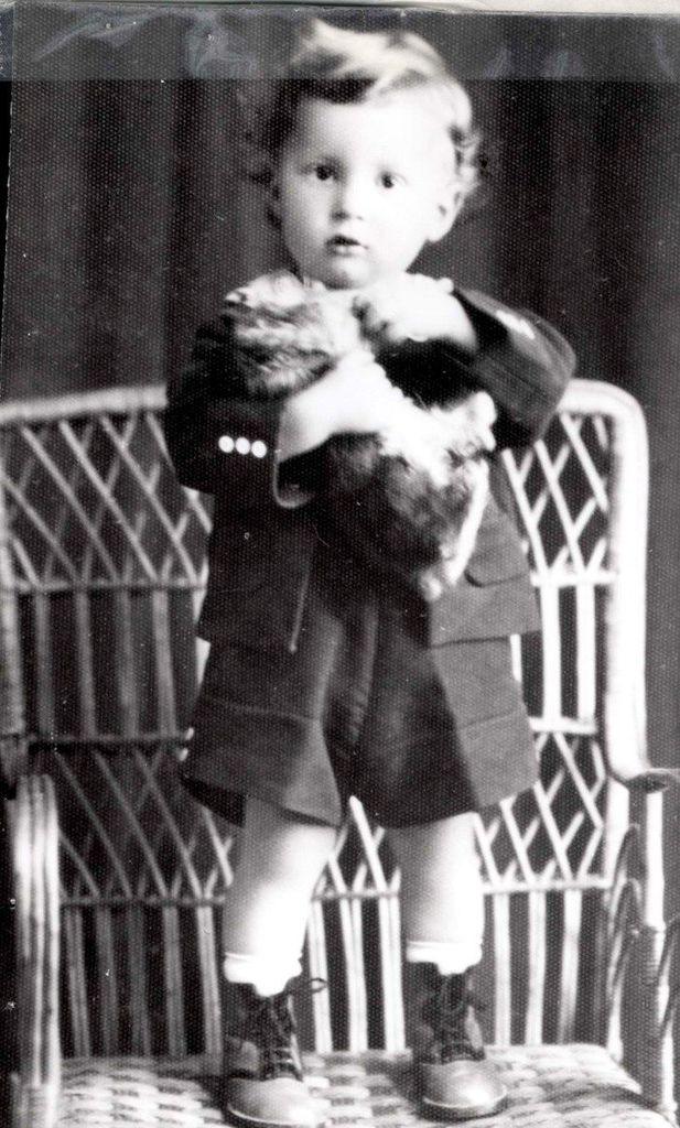 A vintage black and white photo of a young boy standing on a chair holding a toy dog. The child is dressed in a buttoned coat, shorts, and high-top shoes. The chair has a woven backing.