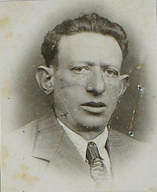 Close-up vintage passport-style photograph of a person wearing a suit and tie, featuring a detailed expression. The photo shows signs of age with slight discoloration and markings.