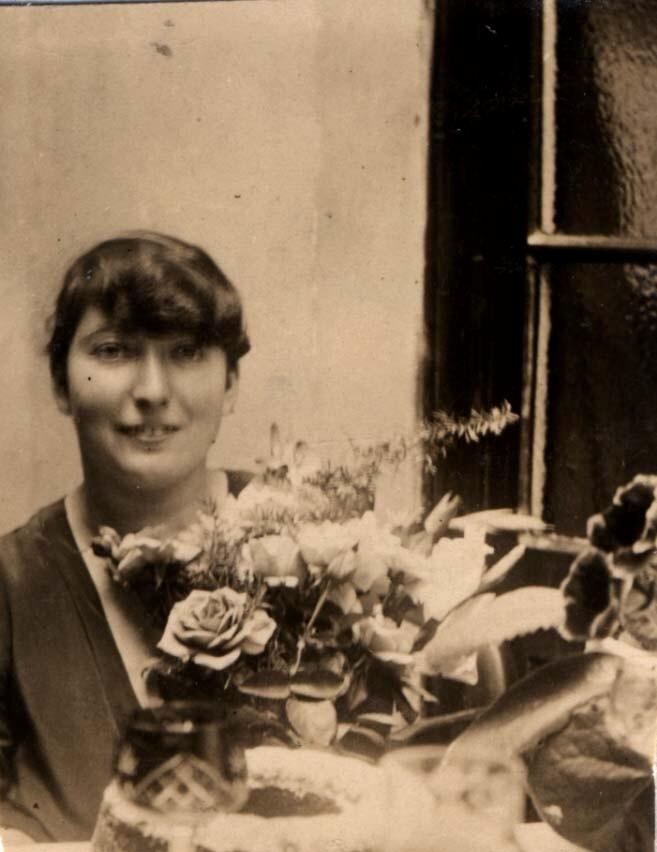 Sepia-toned vintage photograph of a smiling woman seated next to a window, surrounded by an array of flowers.