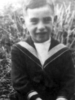 A vintage black and white photo of a young boy smiling in a sailor suit amidst tall grass.
