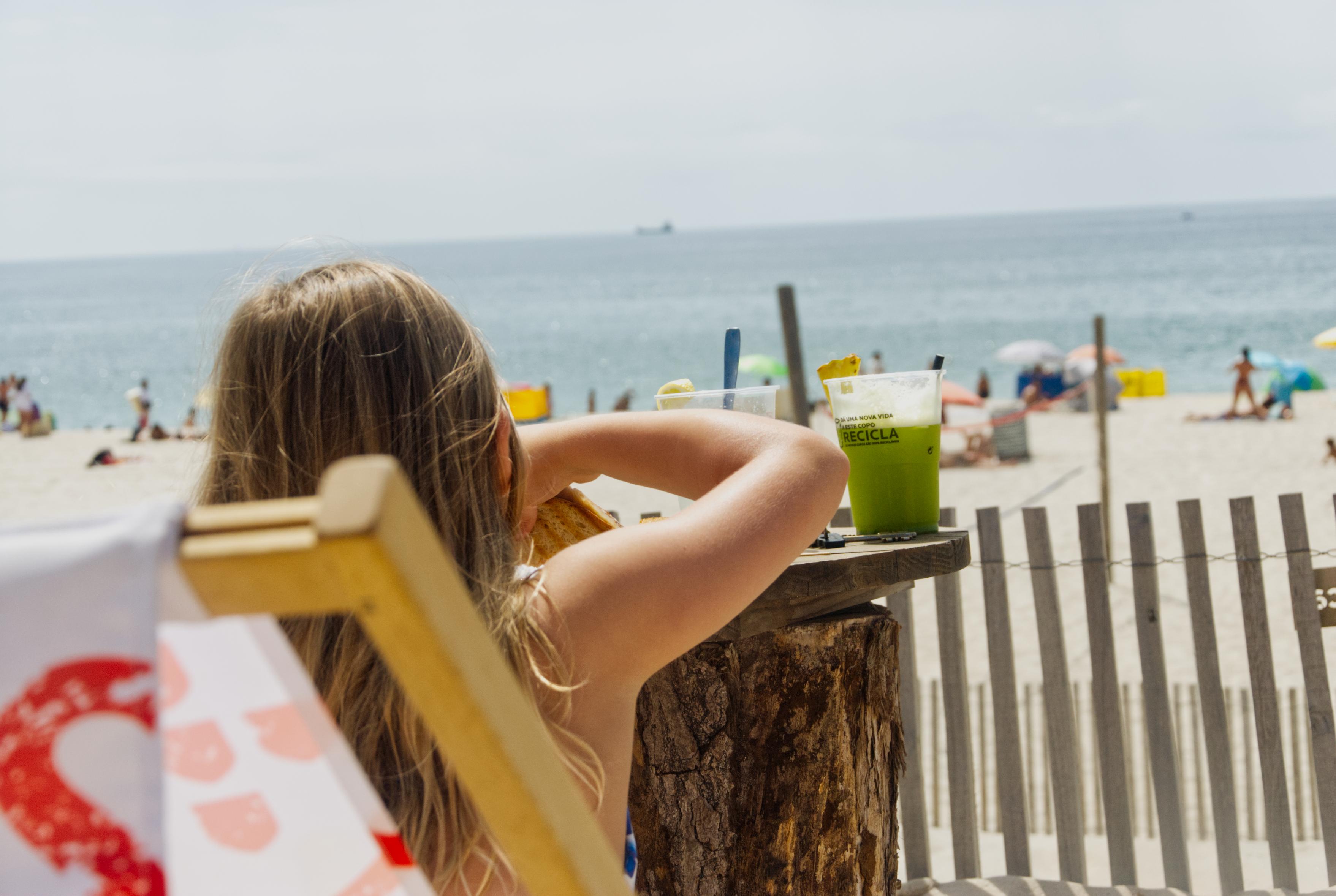 picture of a little girl sitting in a beach chair, back to camera.
she is at a beach cafe, eating, a green drink is at her right elbow.
there is a sand fence, beach, with umbrellas and people, then the water.