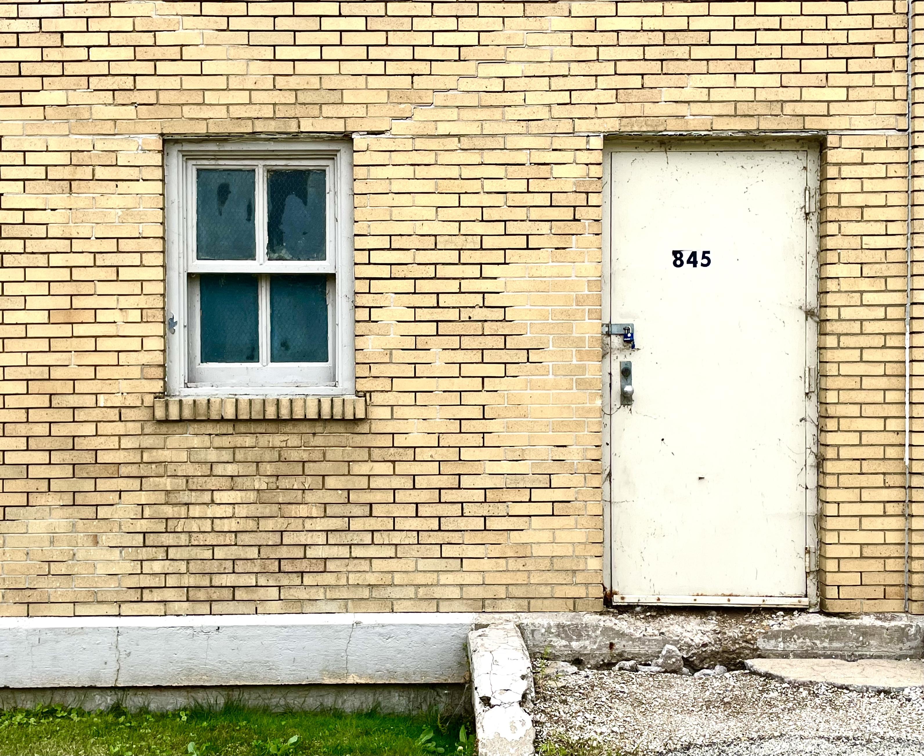 a simple 4 pane window & it's worn white frame beside a plain painted metal door. the wall is yellow brick.  there's the number 845 on the door.