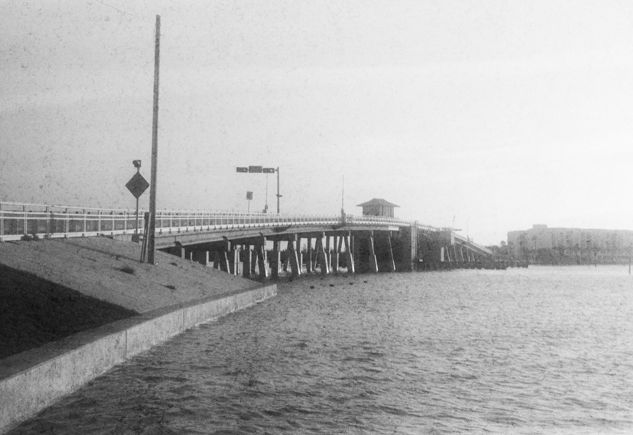 Black and white photograph of a bridge over a fairly large amount of water. The near end of the bridge is to the left of the frame. The bridge is fairly long and the far end of the bridge is near the right side of the frame. There is a small structure in the center of the bridge, and there are some poles, signs, and a traffic light above the bridge. In the distance and in the right side of the frame is a building. The water is mostly still, but ripples can be seen on the surface. The sky appears to be clear. The photograph is rather grainy and low in contrast, as if it were a very old photograph.