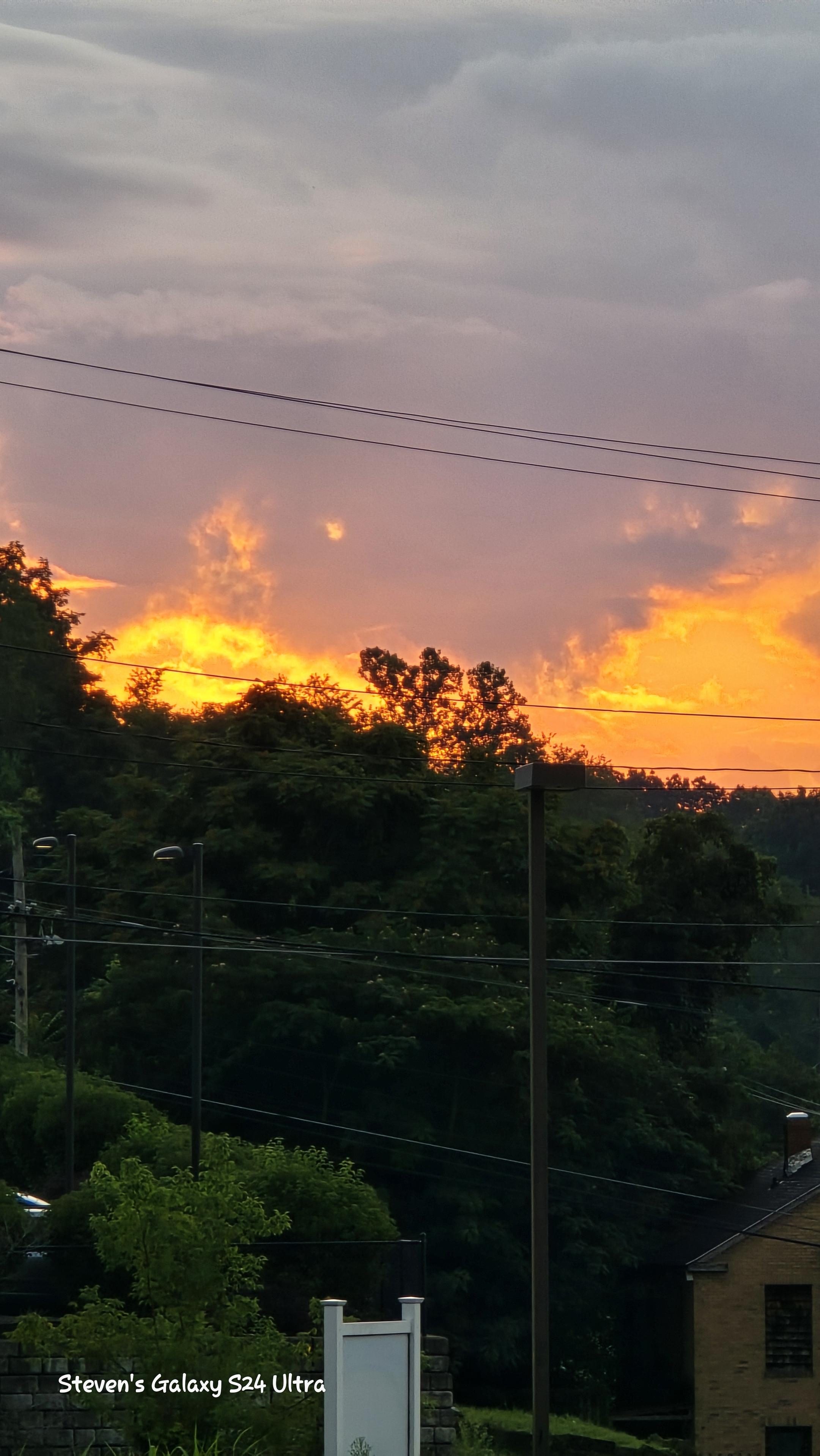 Sunset over lush greenery with vibrant orange and yellow hues in the sky. Power lines cross the scene, adding contrast to the serene landscape.