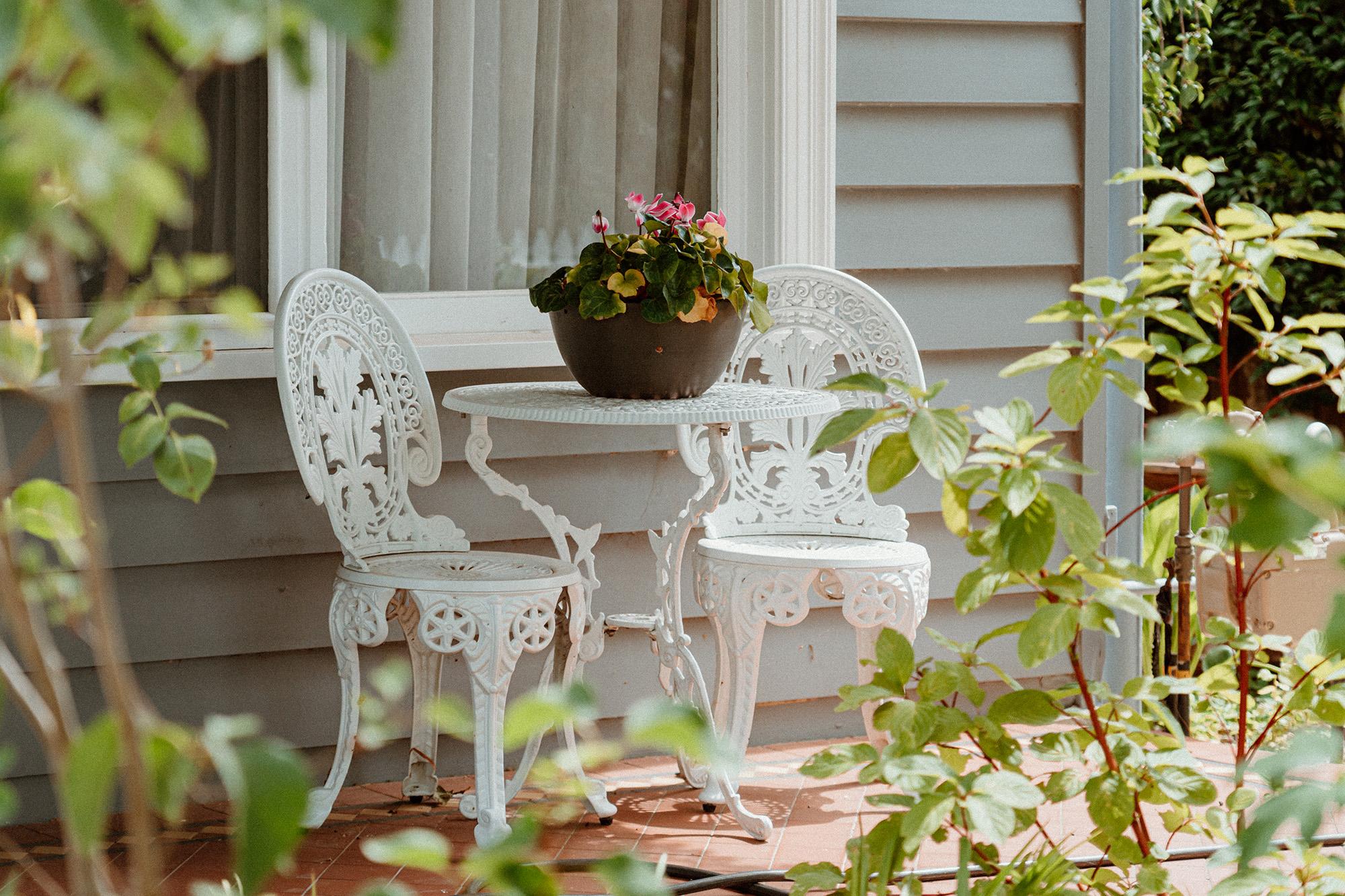 photo of a front porch in Williamstown, Melbourne, Australia. a white cast-iron bistro set on the verandah and a pot of flowers on the table. leaves frame the shot in the foreground. the chairs are set as if waiting for someone coming out with tea. a gentle nostalgic warmth to the image, a bit timeless