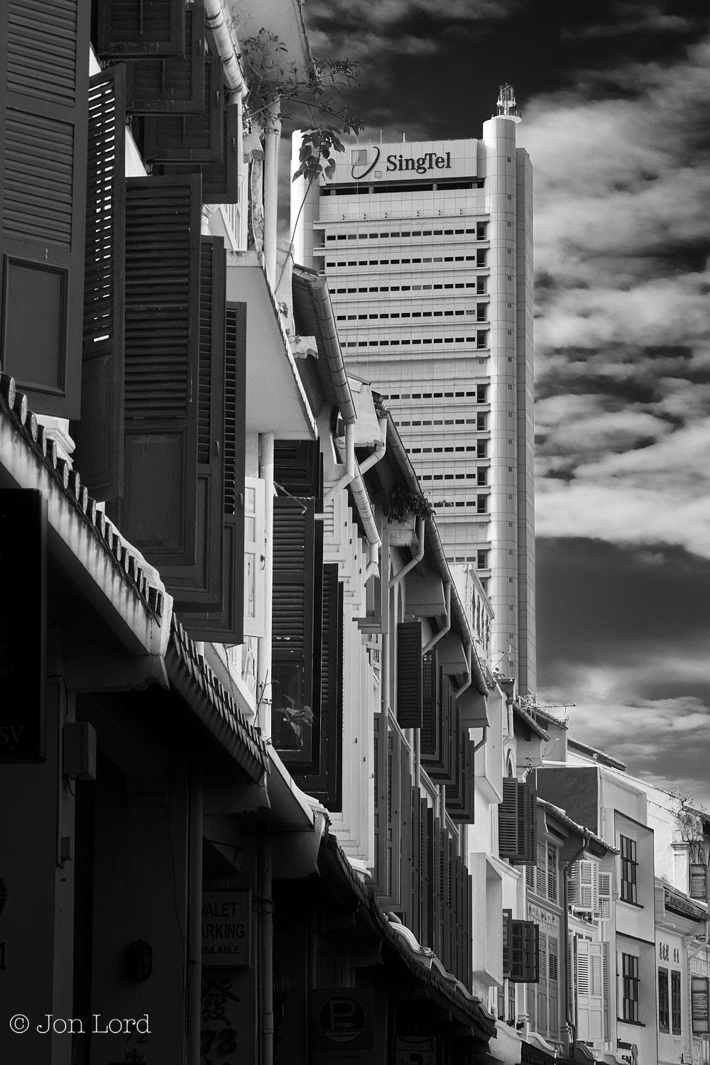 This is a cityscape photo in portrait format of a long row of historic, colonial-era terraced buildings in the foreground and a modern high-rise office block in the background. Amoy Street, Singapore (2012).

In the foreground is that row of old but restored colonial-era terraced buildings that start along the left edge of the photo and extend away from the camera for ten to fifteen metres before curving around to the right and then out of view. The location is a quiet and pleasant area of the city with mostly restored historic low- rise buildings, some residential, but mostly businesses and cafes. The camera is positioned about a metre to the right of the buildings (standing in the road) and aligned viewing along it's length. The buildings are a mix of two and three floors with the ground floor deep in shadow and mostly out of view with what is the pavement covered by the overhang from the first floor of the building, as was so common in that era to protect pedestrians from the tropic