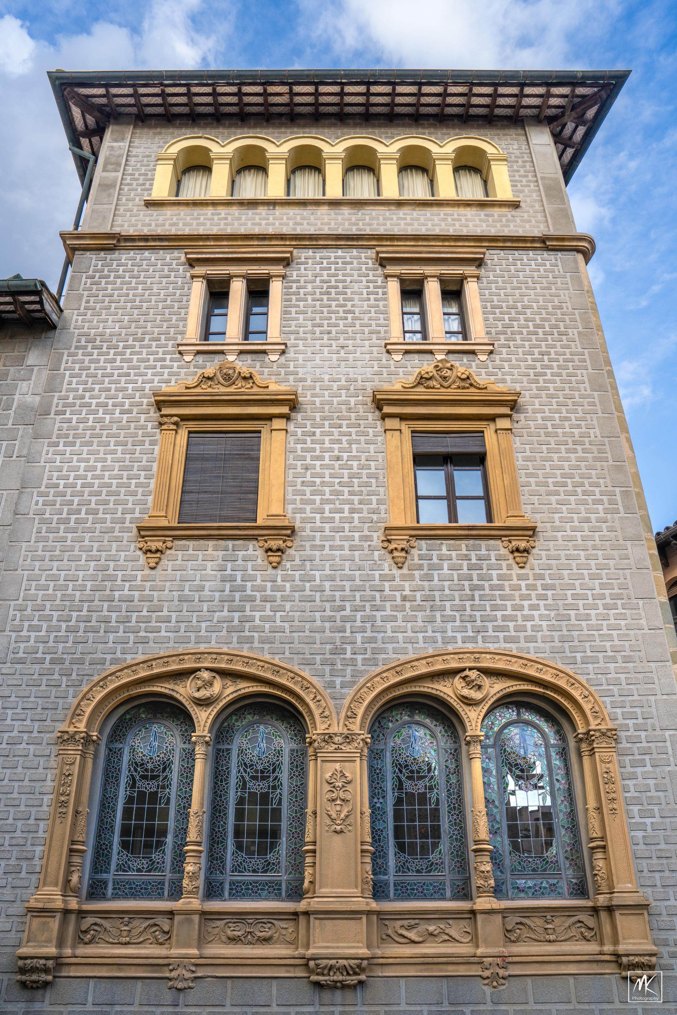 Color photo looking up the facade of a multi-story building tower that has an eclectic mix of classical, baroque, and modernisme elements including elaborately decorated stained glass windows on the lower level.