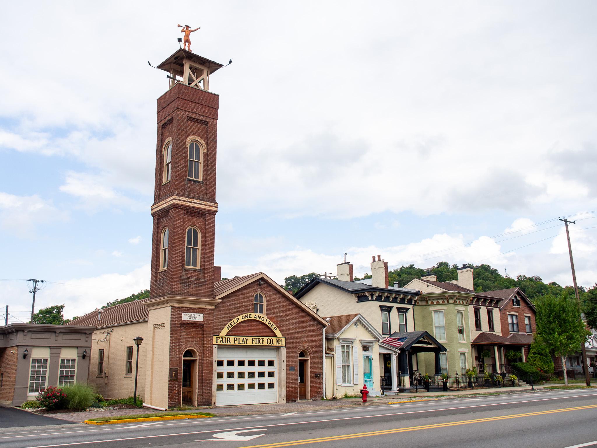 A row of buildings apparently built in the late 1800s, different colors, with ornate trim. Some of the buildings have front porches. At the left end of these buildings is an old red brick firehouse with a tall tower. At the top of the tower is a golden statue of a human figure wearing a hat and blowing a horn. The sky is mostly cloudy.