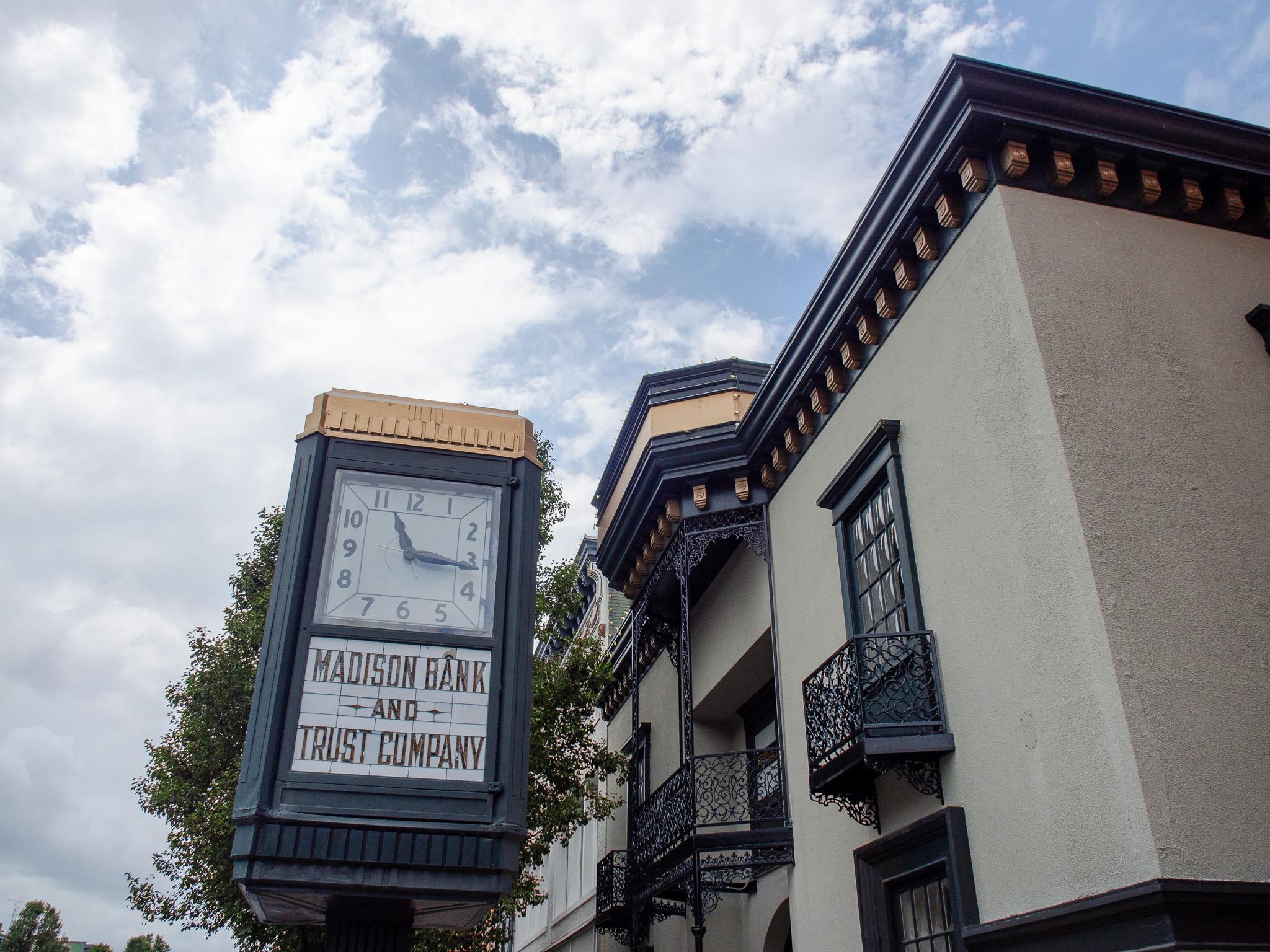 Dominating the right half of the frame is a building shot from below, looking up. It is beige with dark trim at the top and around the windows, with some ornate gold accents. There is some ornate black ironwork around the windows. To the left is a clock mounted on a pole. It is rectangular, painted dark, with some ornate gold trim at the top. Below the clock is a sign that says, "MADISON BANK AND TRUST COMPANY". Behind the clock is a tree with green leaves. The sky is mostly cloudy, with blue space between the clouds.