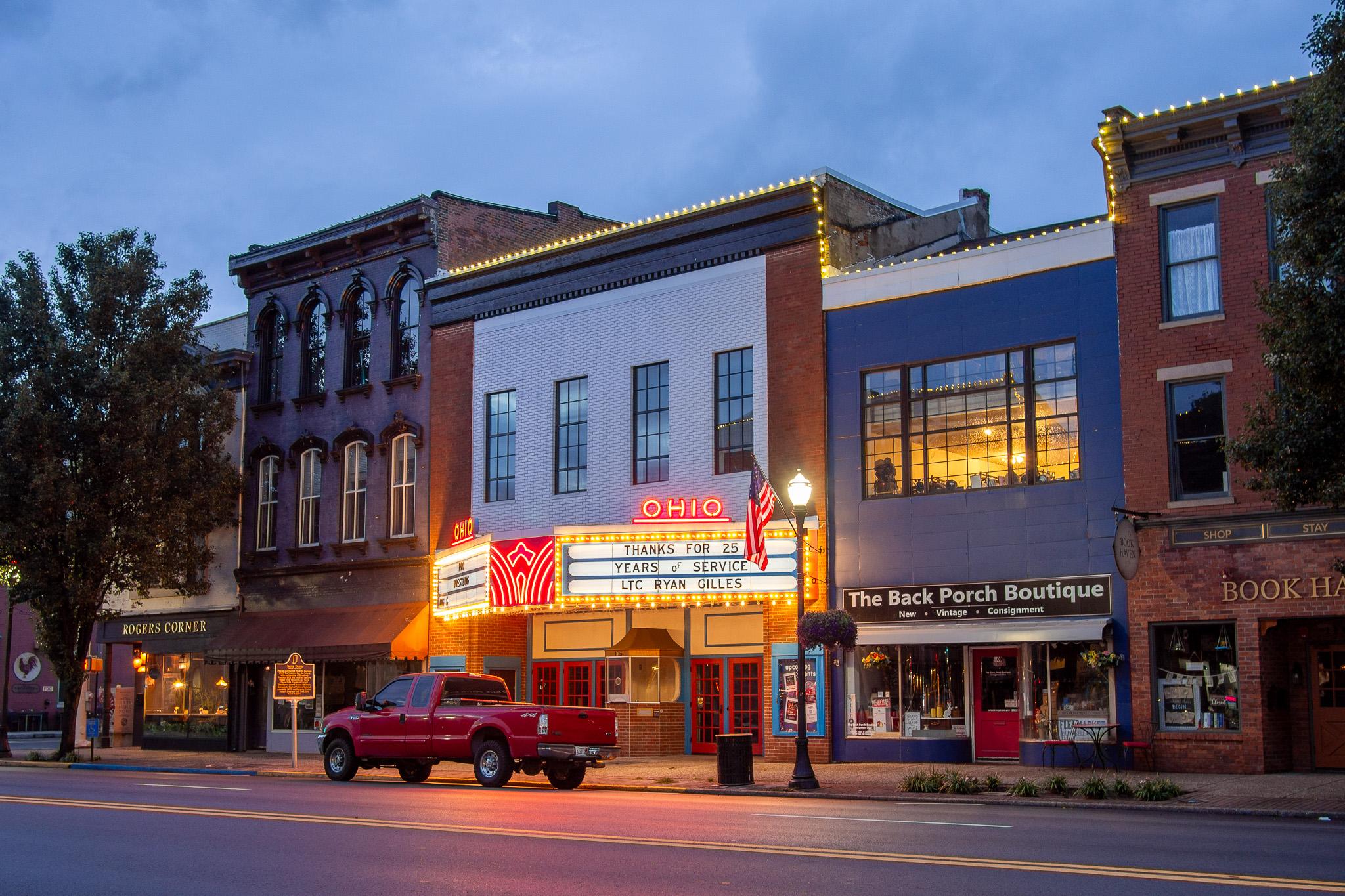 Several buildings in the style typical of 1800s Midwest small towns. The are side-by-side, close to a street, and with a sidewalk in front. The buildings have many windows and storefronts on the ground floor. This is an evening shot, and it is getting dark. There is a string of closely spaced lights along the rooftops. There are lights in some of the windows. The building in the middle of the frame appears to be a theater with a well lit marquee in the front. It says "OHIO" in neon lights at the top. The marquee says "Thanks for 25 years of service. LTC Ryan Gilles". There is a lamp post with a glass lamp shade at the top and an American flag mounted near the top. A red pickup truck is apparently parked in front of the theater. There is a tree in the left of the frame. The sky is dark blue with some clouds.