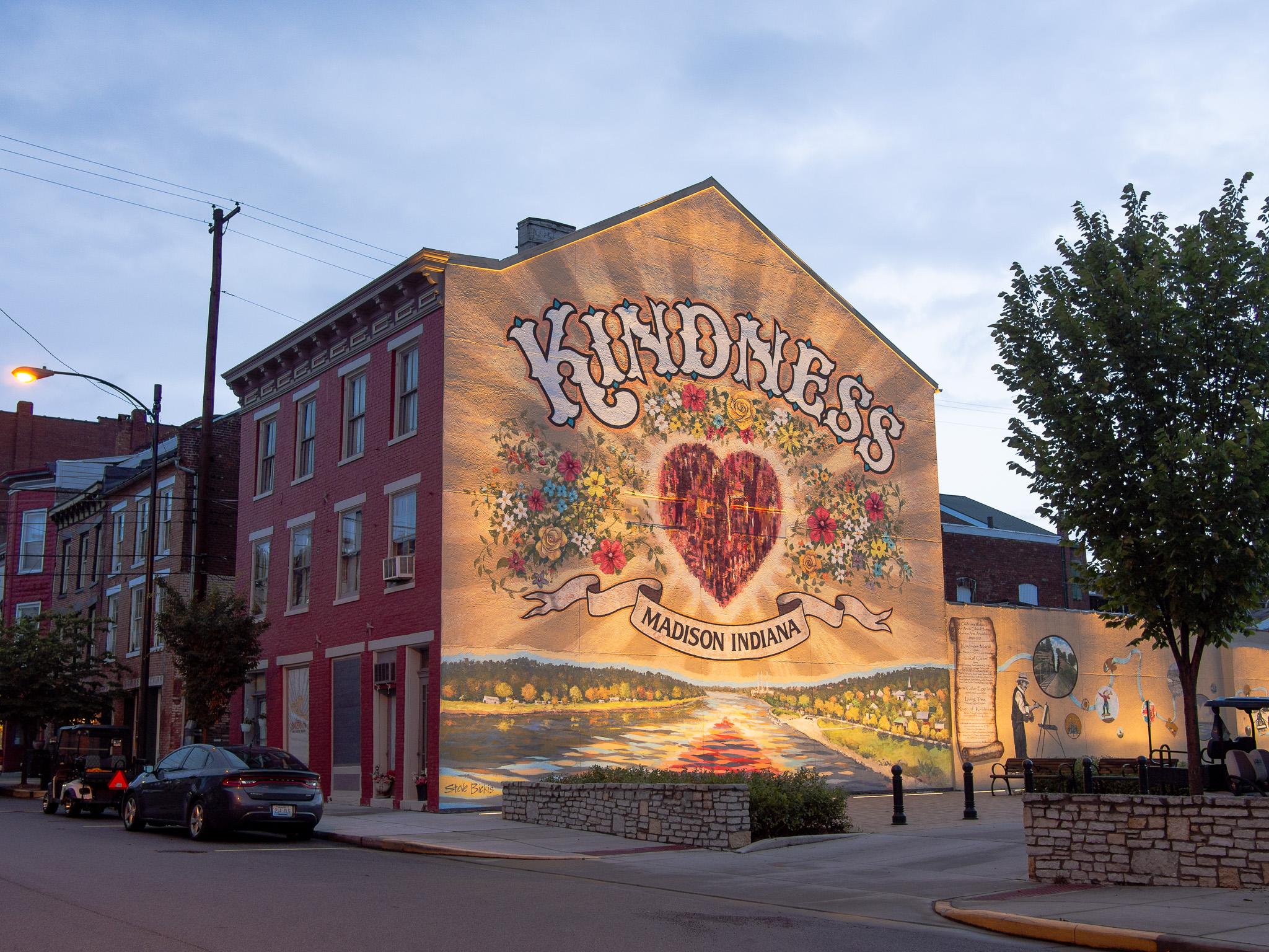 A building with a large mural painted on the side. The mural has the word "KINDNESS' in large white letters at the top. Below that are flower surrounding a red heart. At the bottom are the words "MADISON INDIANA" on a painted floating banner. Below that is some kind of representation of a landscape with a river. In the background of the mural are golden rays radiating out from the center. The photo was taken in the evening in partial darkness, and the mural is well lit. To the left are other buildings in the background and some cars parked on the street. To the right are some brick walls and a tree with green leaves. The sky is dark blue with thin white clouds throughout.