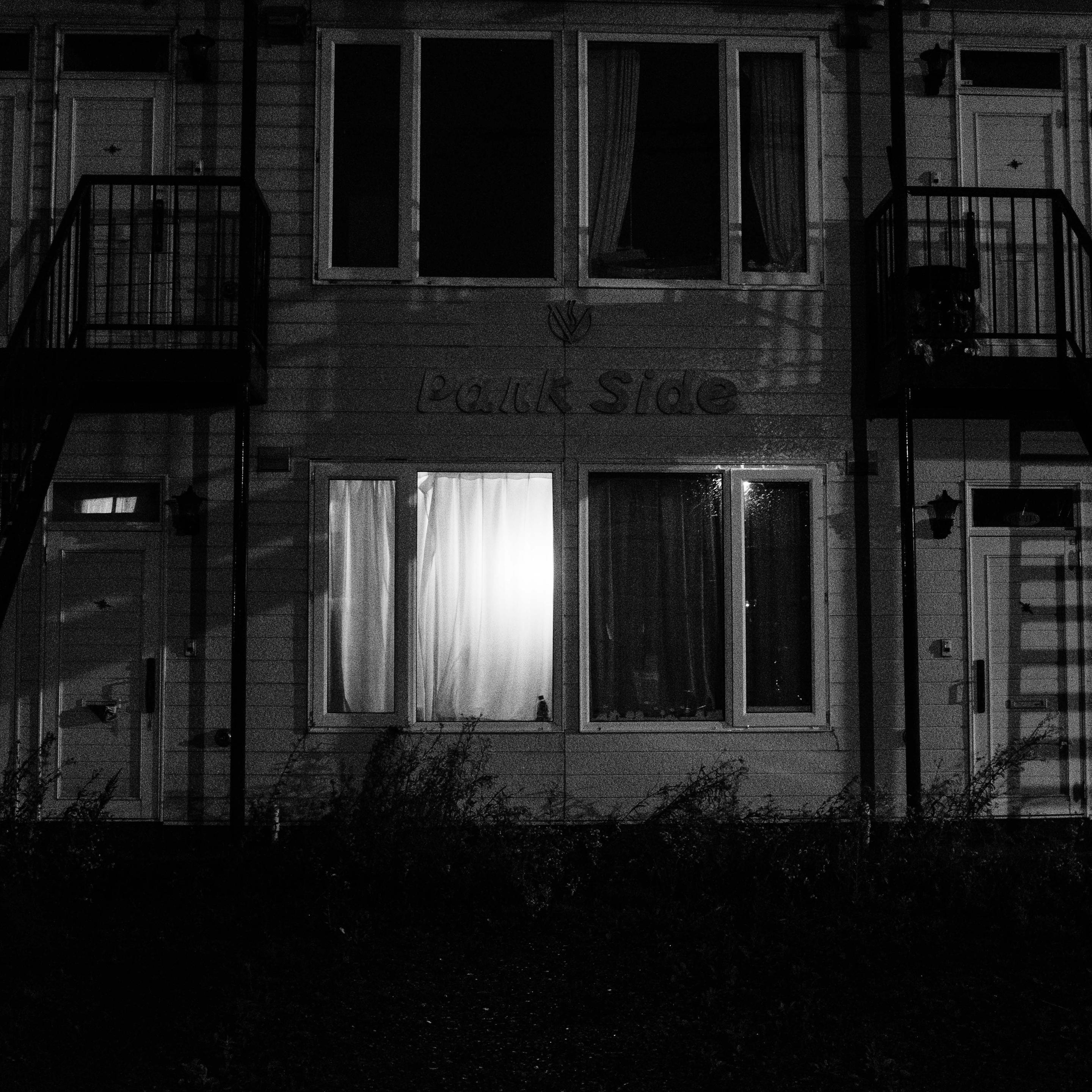 A monochrome photograph captures the front of a two-story apartment building at night. Only one ground-floor window in the center is brightly lit from within, shining through a white curtain. Other windows and doors to the sides and above are dark. Metal stairs and balconies are visible.