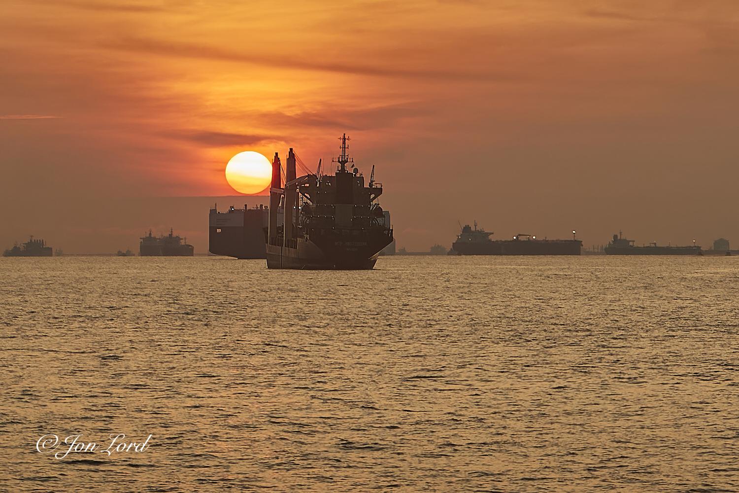This is a colourful seascape photo in landscape format of the anchorage in the Singapore Strait with multiple anchored cargo ships together with the morning sun only a few degrees above the horizon. Singapore (2017).

The relatively calm sea with only small wavelets disturbing the surface, rises upwards from the base of the photo to the halfway up level where there is a crisp horizon dividing the sea from the sky. In the centre of the photo is the stern of a medium sized mixed cargo ship, at anchor. The view is slightly oblique with the stern and port side in view and at a distance of about five hundred metres and with the height of the ship being about a quarter of the height of the image. The ship is partially in shadow though the stern superstructure is clearly defined with multiple bright lights illuminating the four visible decks and with the wheelhouse on top. On the port side are two large cargo handling cranes. The ship is angled bow up slightly suggesting the cargo holds are e