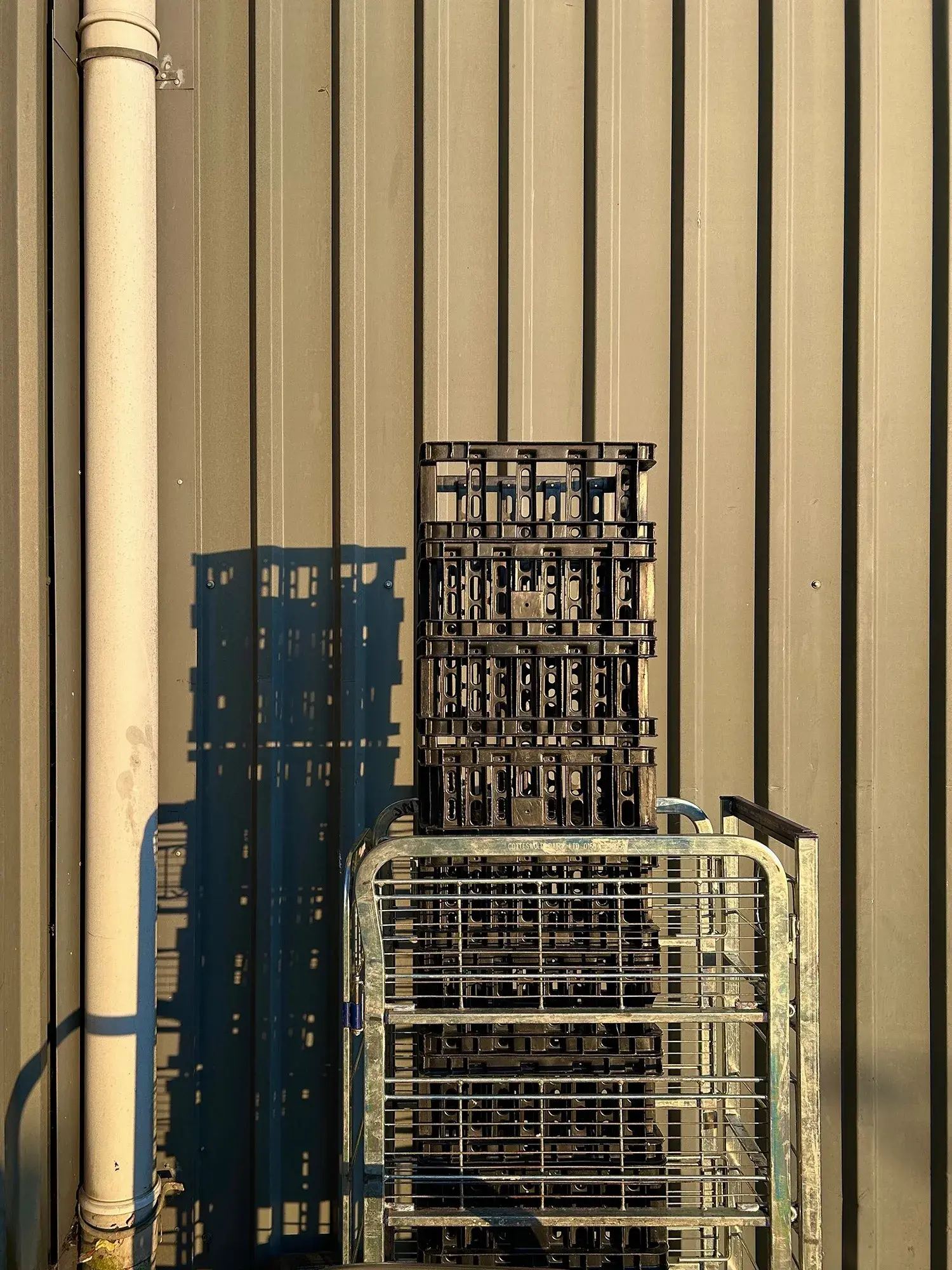 Exterior photograph of some plastic crates stacked within a rolling cage. The dawn light renders the scene in a golden glow and casts a sharp, intricate shadow on the corrugated metal background. Newark Works, Foundry Lane, Bath, UK. Photo by Gearóid Burke.