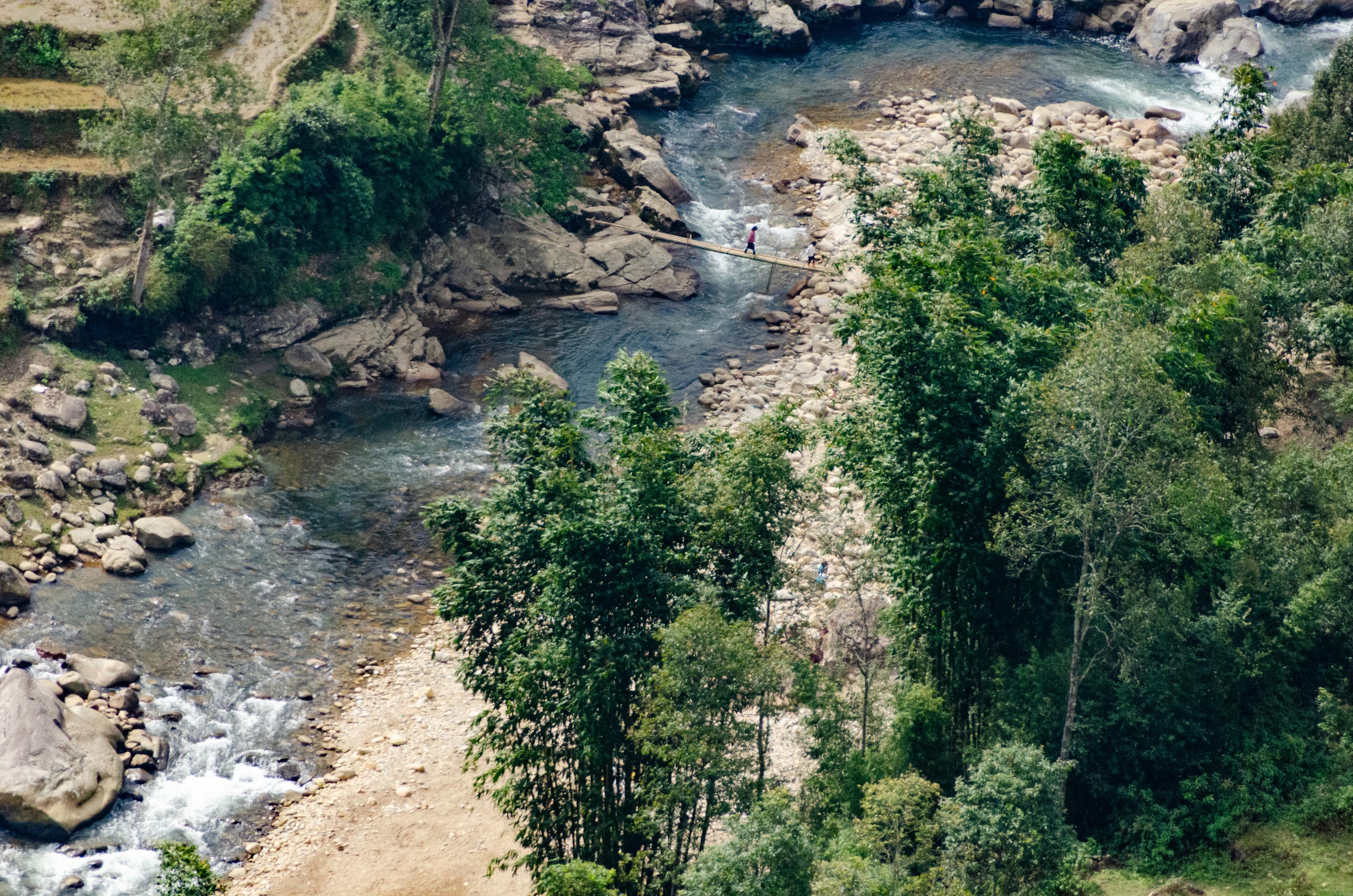 a photo of a river gorge seen from high above, somewhere in Sapa, Vietnam. terraced rice fields on the upper left, a winding mountain river with milky blue-grewen water and a tiny figure crossing a narrow footbridge in the center of the frame.