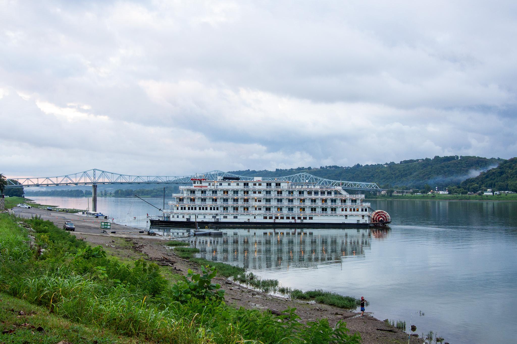 Large riverboat with multiple levels and many doors, apparently a  commercial cruise boat, with a red and white paddle wheel on the back. The boat is mostly painted white. It is sitting on a large river with still water. Behind the boat is a large bridge that crosses the river. In the foreground is grass and an area of sand or dirt close to the river. There are some cars near the river in the distance. The far bank of the river is very hilly and green with leafy trees. There is a bit of fog between the hills on the far right. The sky is dark with heavy clouds. There is a bit of pink on the horizon. There is a blue tint to the entire scene.