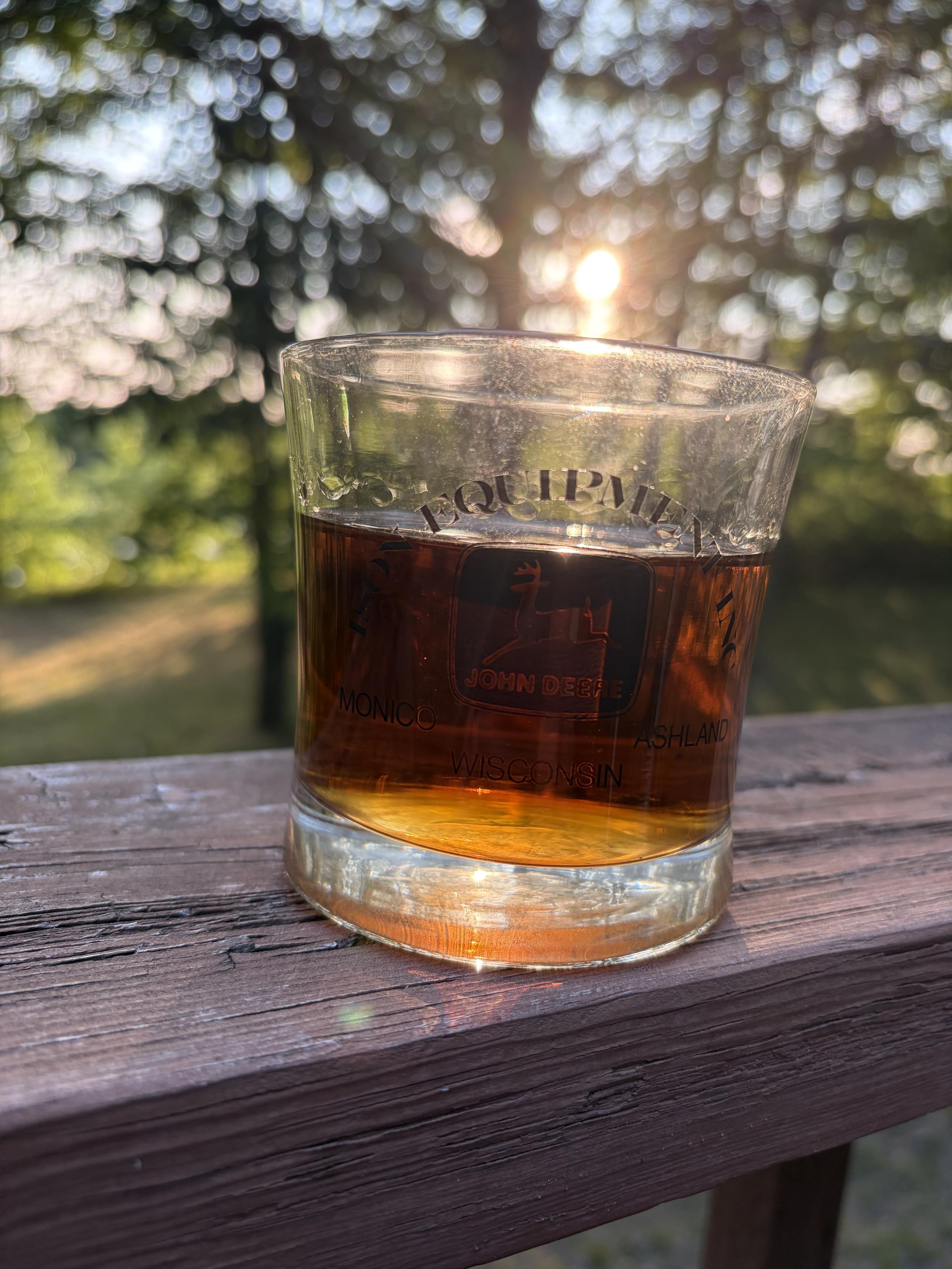 A photo with a glass of bourbon sitting on a deck rail with the sunset behind it.