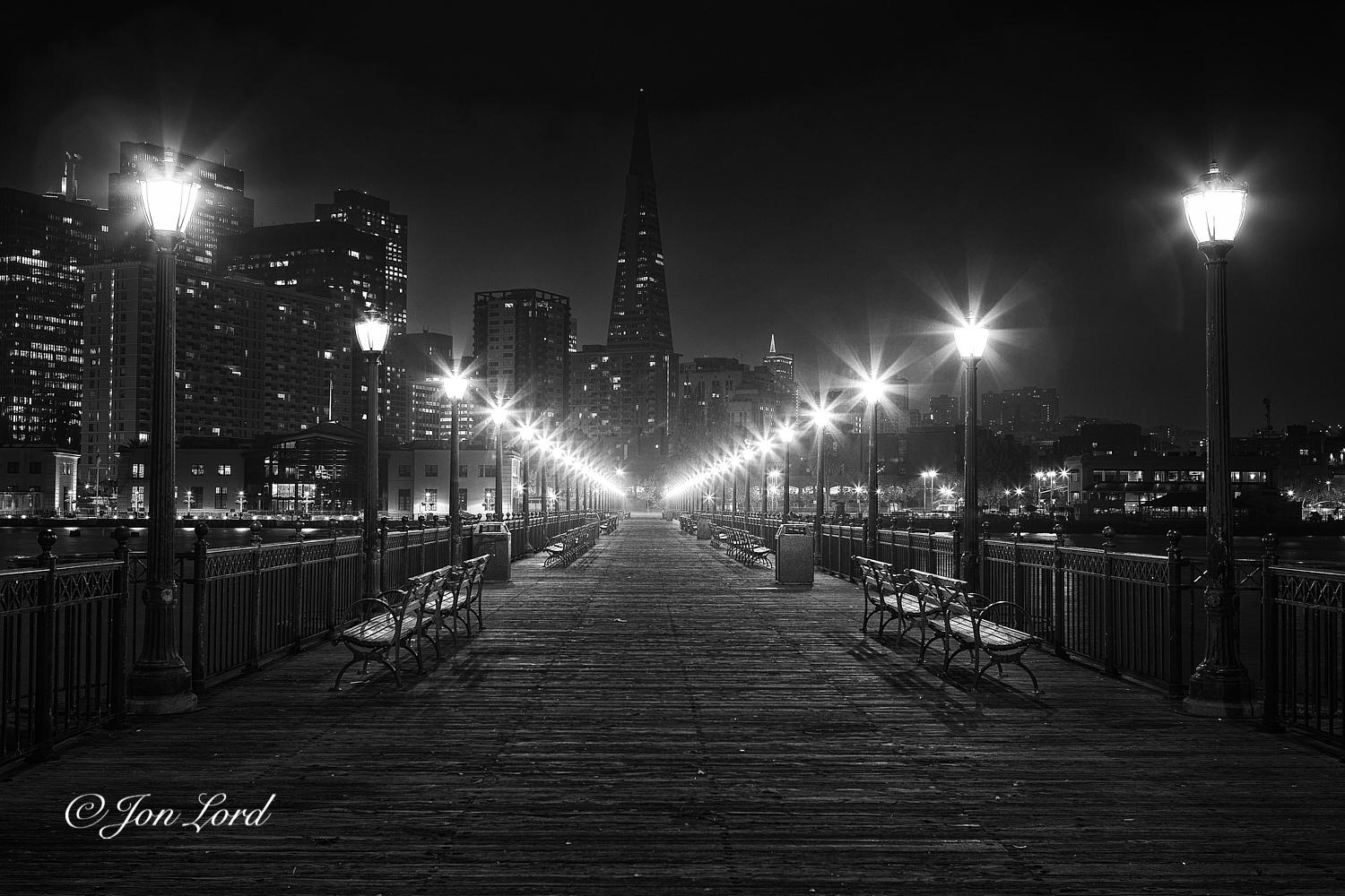 This is a grungy night time photo looking along a wooden pier aligned with and  towards the TransAmerica Pyramid in downtown San Francisco (2012).

Extending away from the camera and into the distance, about 150m away is the wooden pier. The rough wooden planks that form the surface cross at right angles to the pier and stretch away from the base of the image to the centre of the photo. On the side of the pier are metal railings with illuminated ornamental street lights spaced every 3 or 4 metres and casting a star like glow or aura around the lamp. Set inwards a metre of so from the railing are pairs of park benches facing outwards. The wooden planks, metal railings and street lights converge to a point with increasing distance towards the city end of the pier. Beyond is a night time cityscape with the high-rise buildings descending downwards from the business district on the left side to the low-rise residential buildings on the right. Aligned with the pier and in the middle ground, 