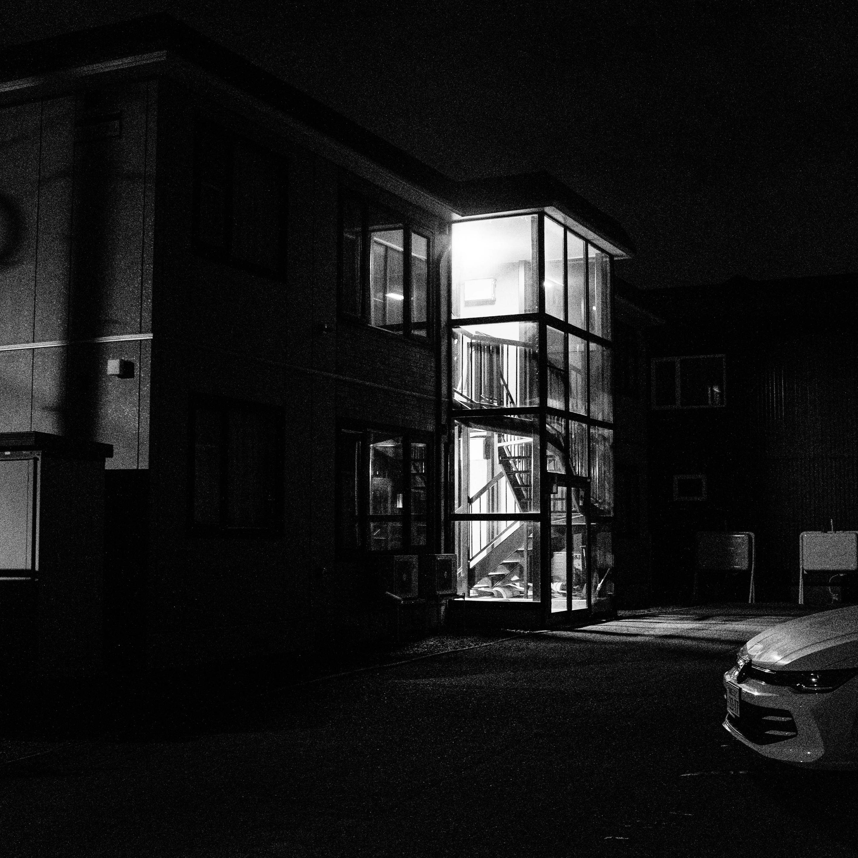 A black-and-white photograph shows a two-story building at night. A central glass-enclosed stairwell is brightly lit from within, revealing an interior staircase. The surrounding facade and parking area are in shadow, with the front of a car visible on the bottom right.