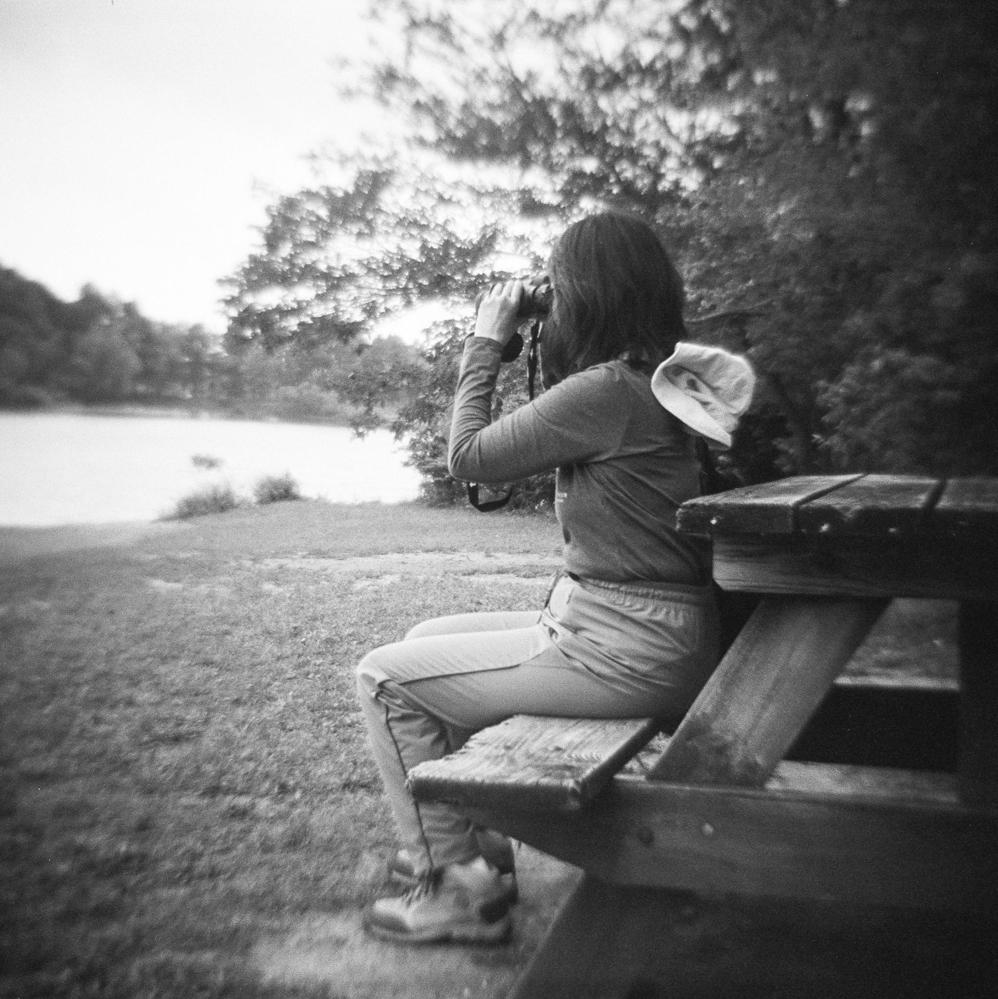 Black and white photo of a woman sitting on a picnic table bench, facing away from the table. She is wearing hiking boots, hiking pants, a long-sleeved shirt, and has a hat hanging behind her from a string around her neck. She has dark, shoulder-length hair, and is looking through a pair of binoculars. The ground is covered with short grass. In the background is a portion of a lake, with trees on the distant shore. Behind the picnic table there are trees filling the frame on the right side. The image is square, and it is sharp in the middle of the frame, but more blurry in the distance and around the edges of the frame.