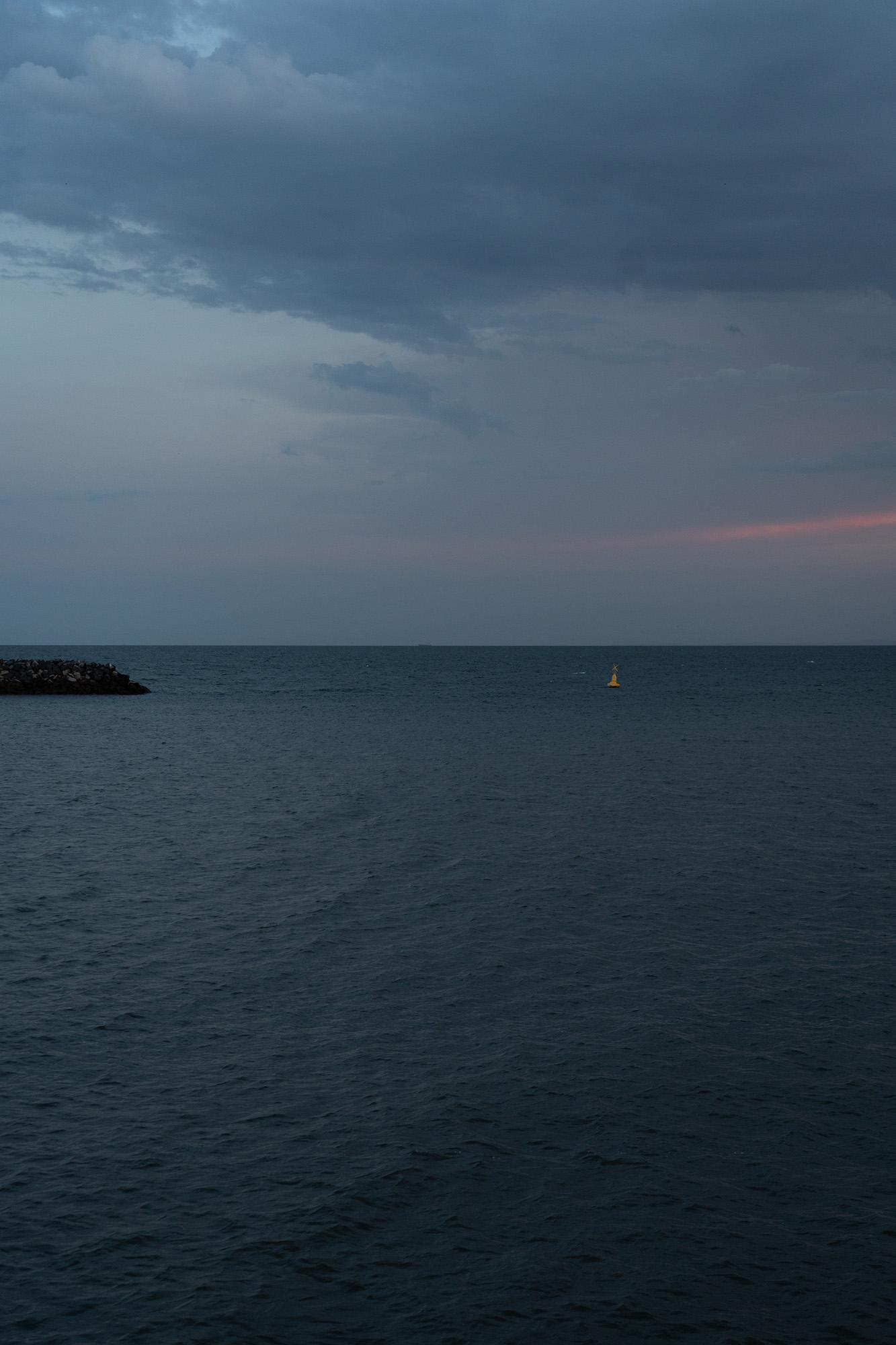 a photo of the ocean at blur hour. dark water, cloudy sky, a thin streak of pink or red in the clouds at the horizon and a rocky breakwater on the left. one small buoy in the distance. taken in Redcliffe, Australia
