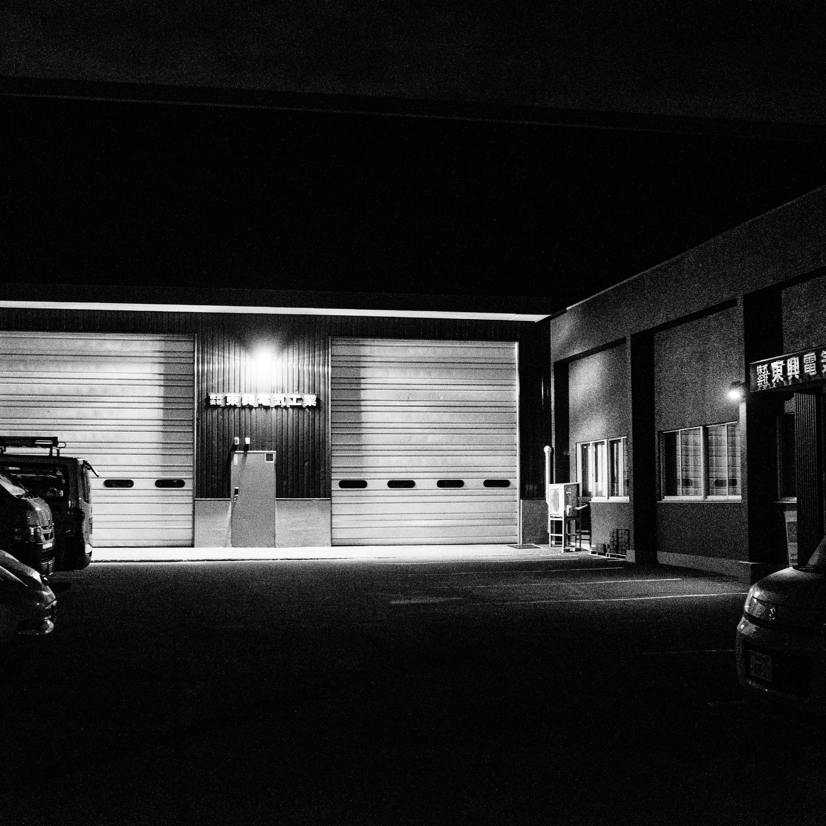 A black-and-white night photograph of an industrial building. Two large closed roll-up shutter doors are illuminated by exterior lights. Vehicles are parked in the dark asphalt lot. Bright light highlights the walls and ground, contrasting with the surrounding darkness.