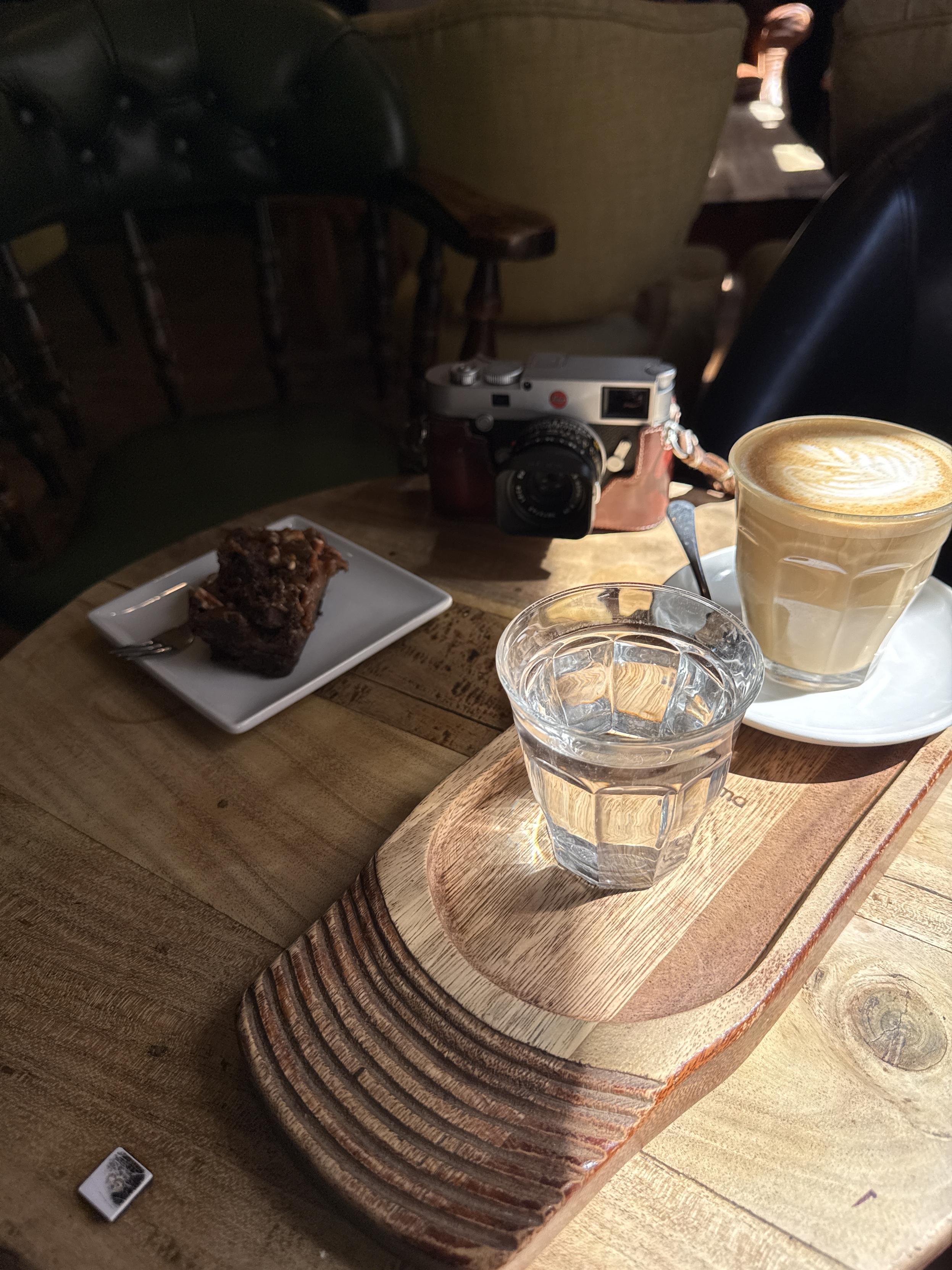 A cozy café setting featuring a wooden table with a latte art coffee in a glass, a small plate with a piece of dessert, a glass of water, and a vintage camera. Soft lighting enhances the warm ambiance.