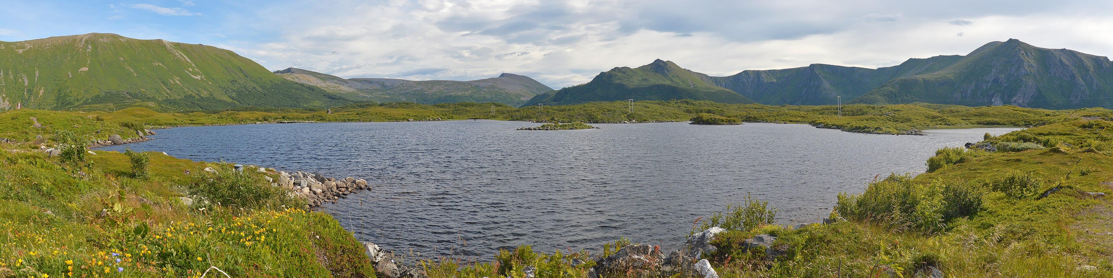 A panoramic photo of a lake surrounded by low growing plants. There are mountains in the distance. The sky is filled with clouds.