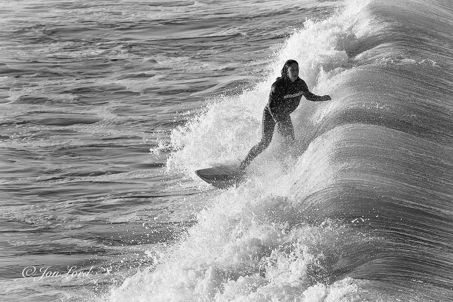This is a black and white water sports photo in landscape format of a female teen surfing shortly after sunrise. Hermosa Beach, California (2013).

Stretching from the right edge of the photo to a little less than the half way across point is a fierce braking Pacific wave. The wave which is now a little less than two metre high, is moving from right to left, viewed from two metres above and a few metres away from the closest point. The wave has multiple streaks or lines extending at right angles to the wave, from the crest and out to sea. Ahead of the breaking wave is our young female surfer. She is positioned slightly to the right of centre in the maelstrom of white foaming surf and is in motion diagonally across the photo from the centre towards the lower left corner. Our surfer is viewed from the front, with her standing on the board sideways (standing on the board and facing to the left). She is viewed from above at five metres distant and with her occupying half the height of the 