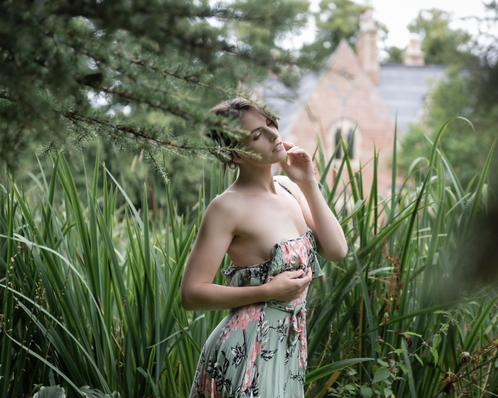 A woman in a floral dress stands among tall greenery, gently touching her chin and looking serene. A house is faintly visible in the background, partially obscured by foliage.