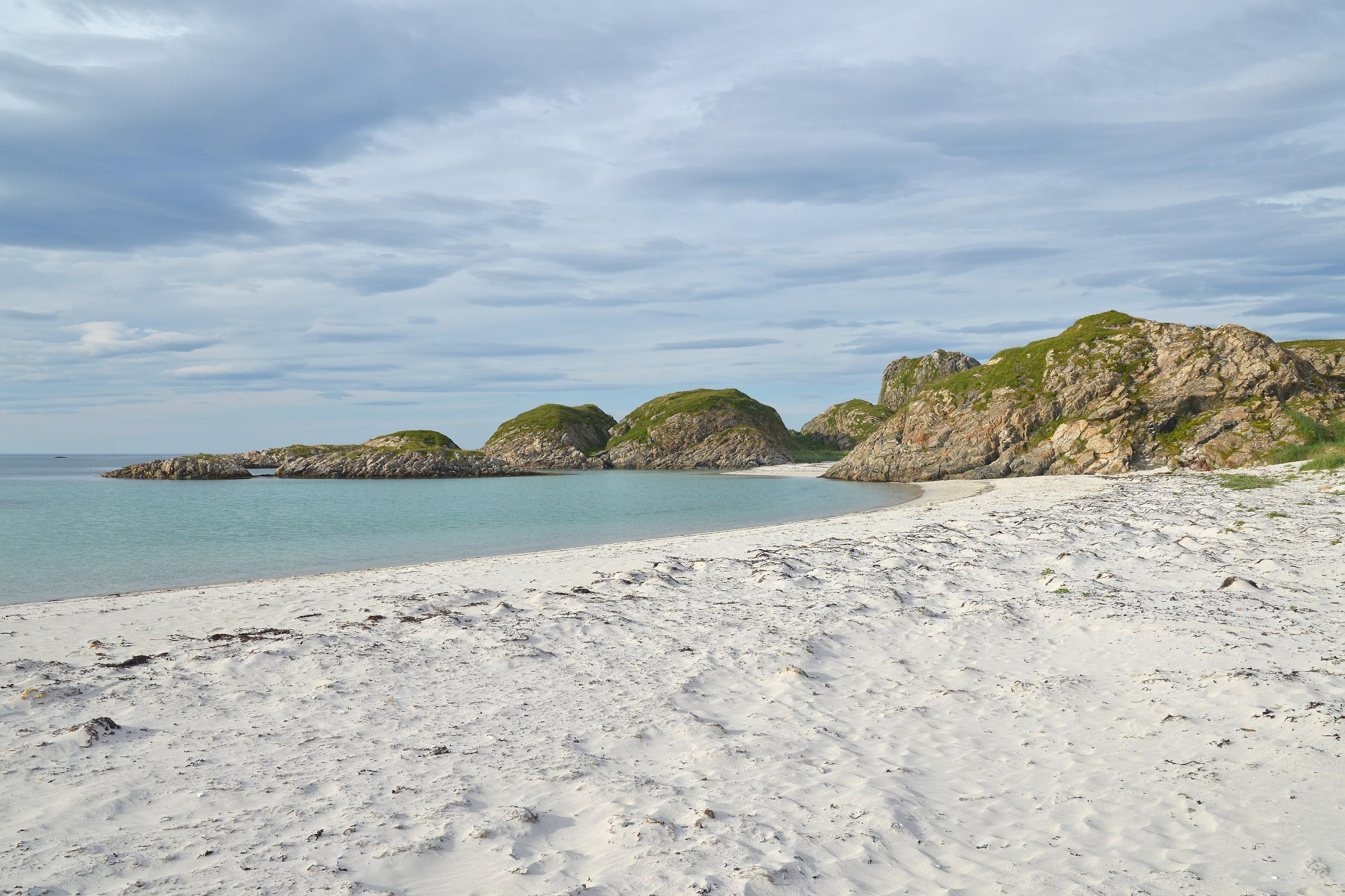 A photo of a sandy beach leading into rocky outcrops. The sky is filled with clouds.