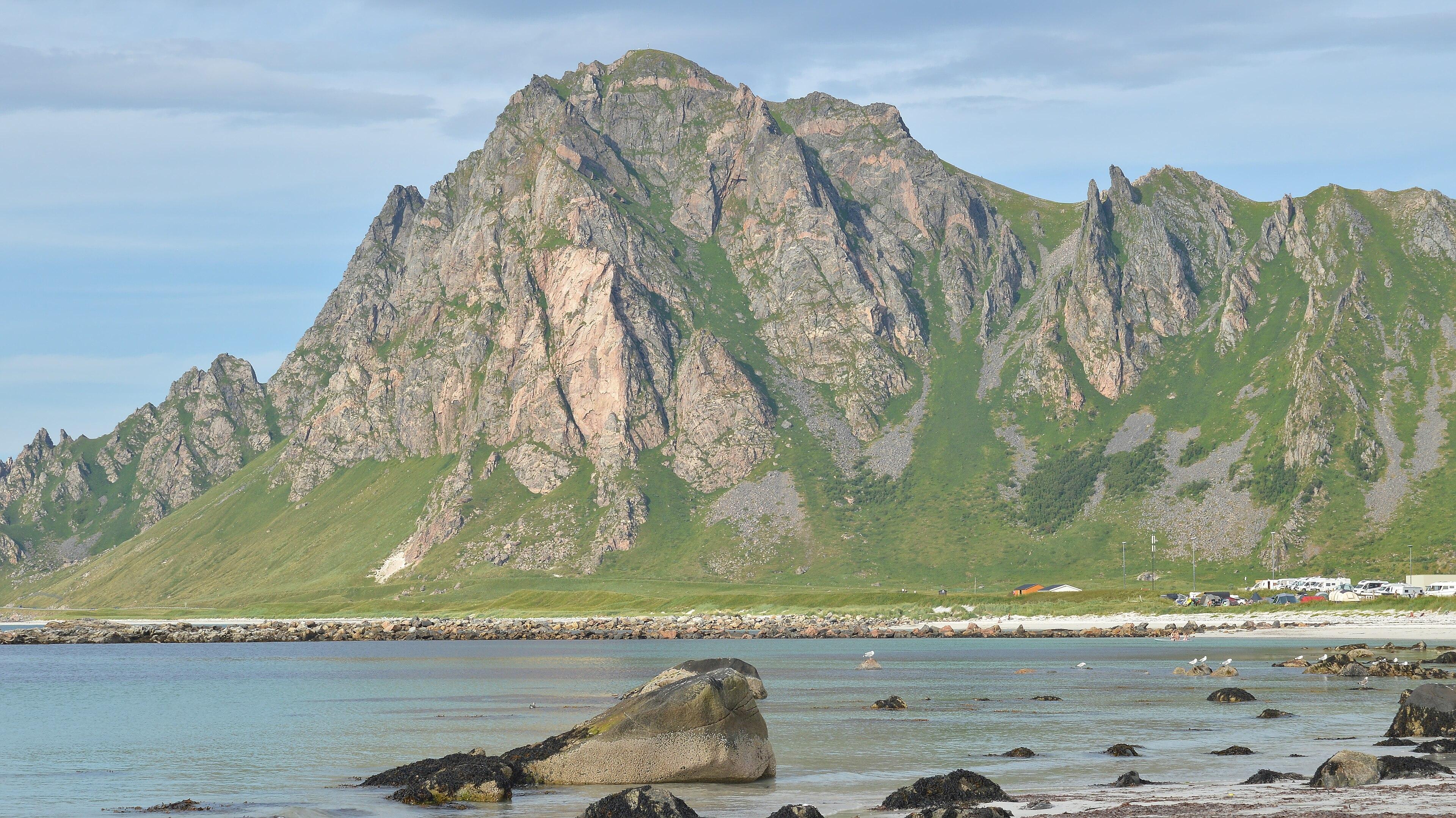 A photo of jagged mountains with water in front of them. The sky is filled with clouds.