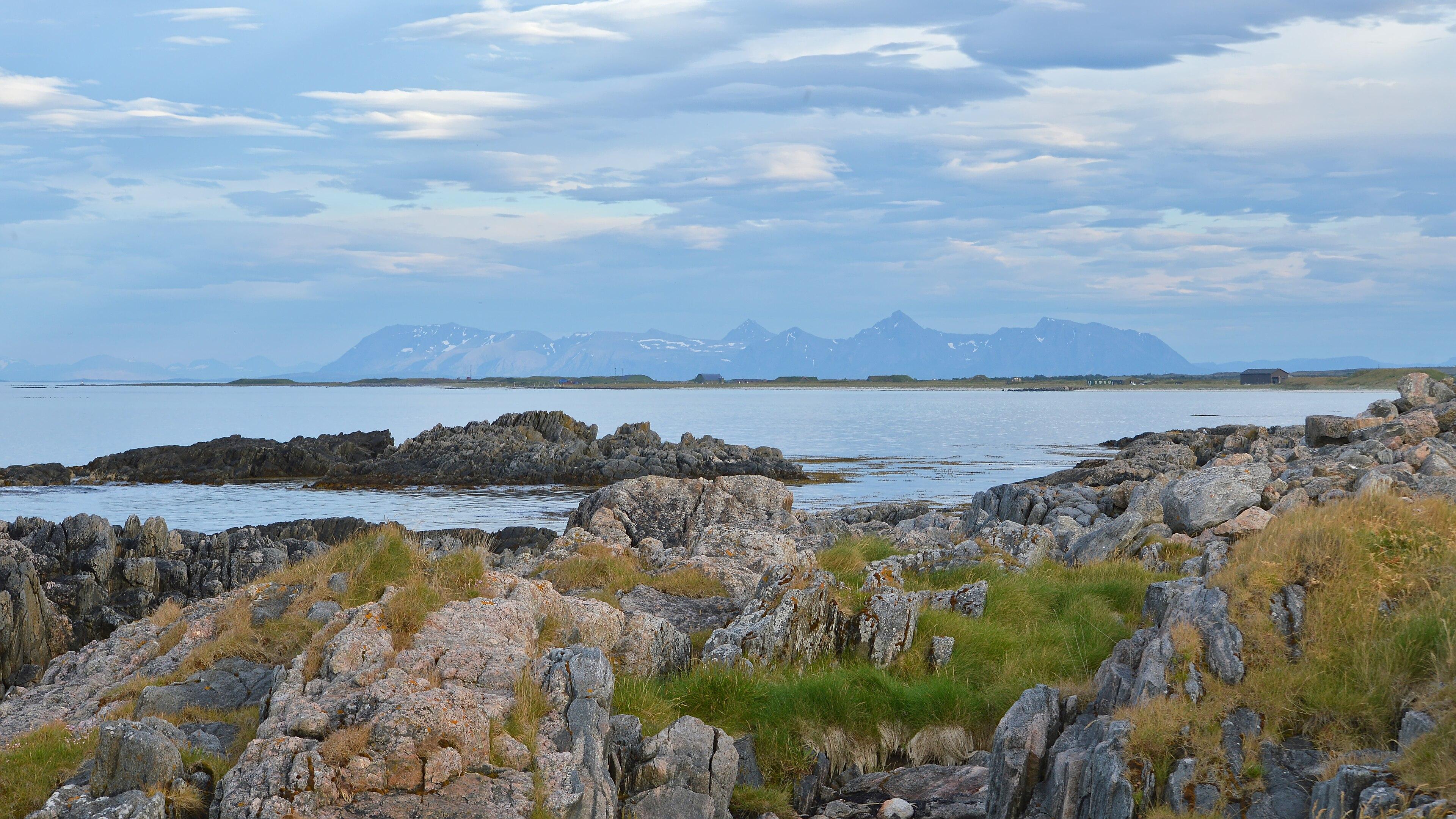 A photo of a rocky shore with a mountainous island in the distance. The sky is filled with clouds.