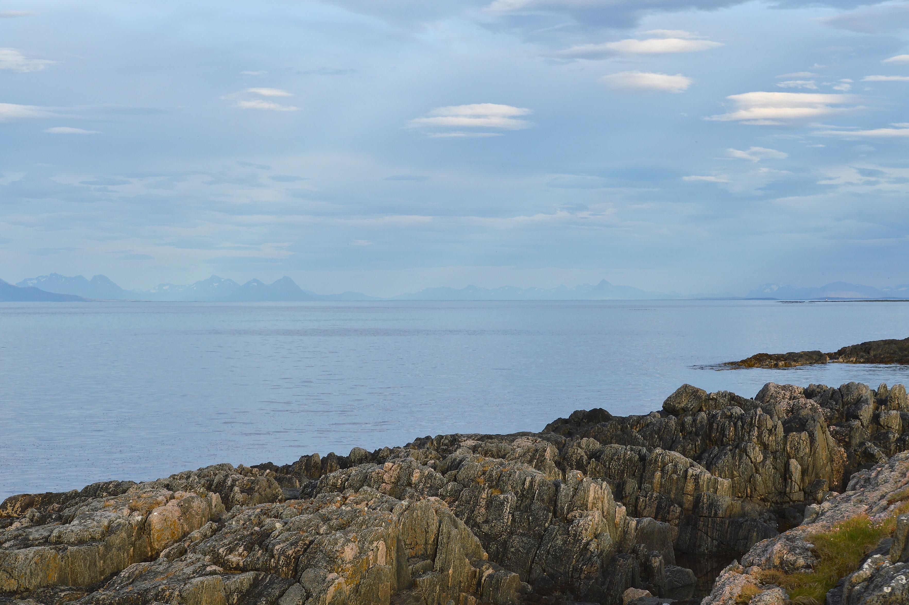 A photo of a fjord with a rocky shore and mountains far in the distance. The sky is filled with clouds with some crepuscular rays shining through.