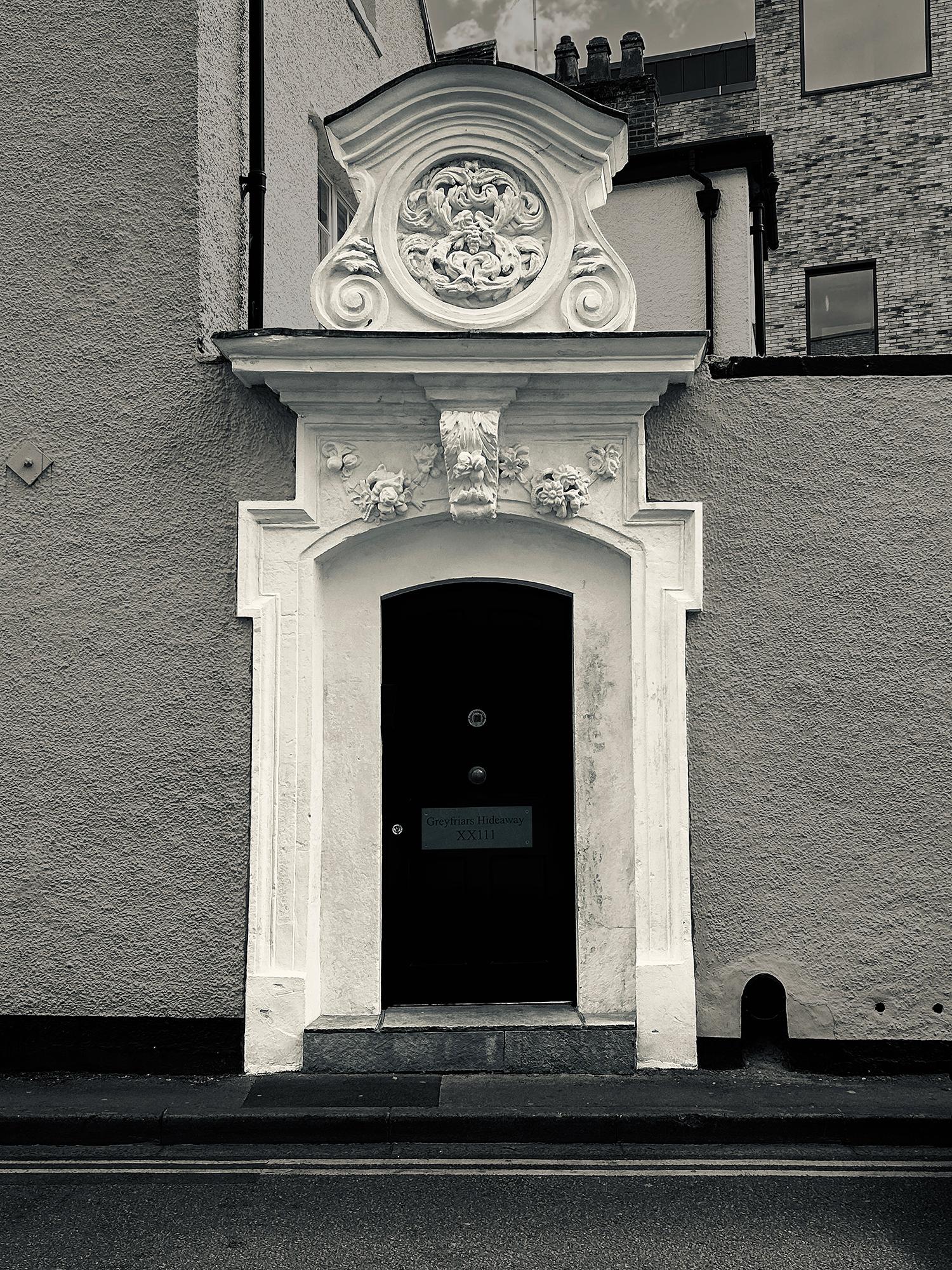 A monotone photograph of a white-washed doorway featuring a prominent, elaborate Baroque-style moulded stucco overdoor pediment. The comparatively basic door is painted black and has a cheap sign that reads "Greyfriars Hideaway XXIII." Perhaps surprisingly, this is the entrance to the courtyard of a grade-II-listed, 17th-century townhouse and "5 Star" luxury holiday rental. Paradise Street, Oxford, UK. Photo by Gearóid Burke.