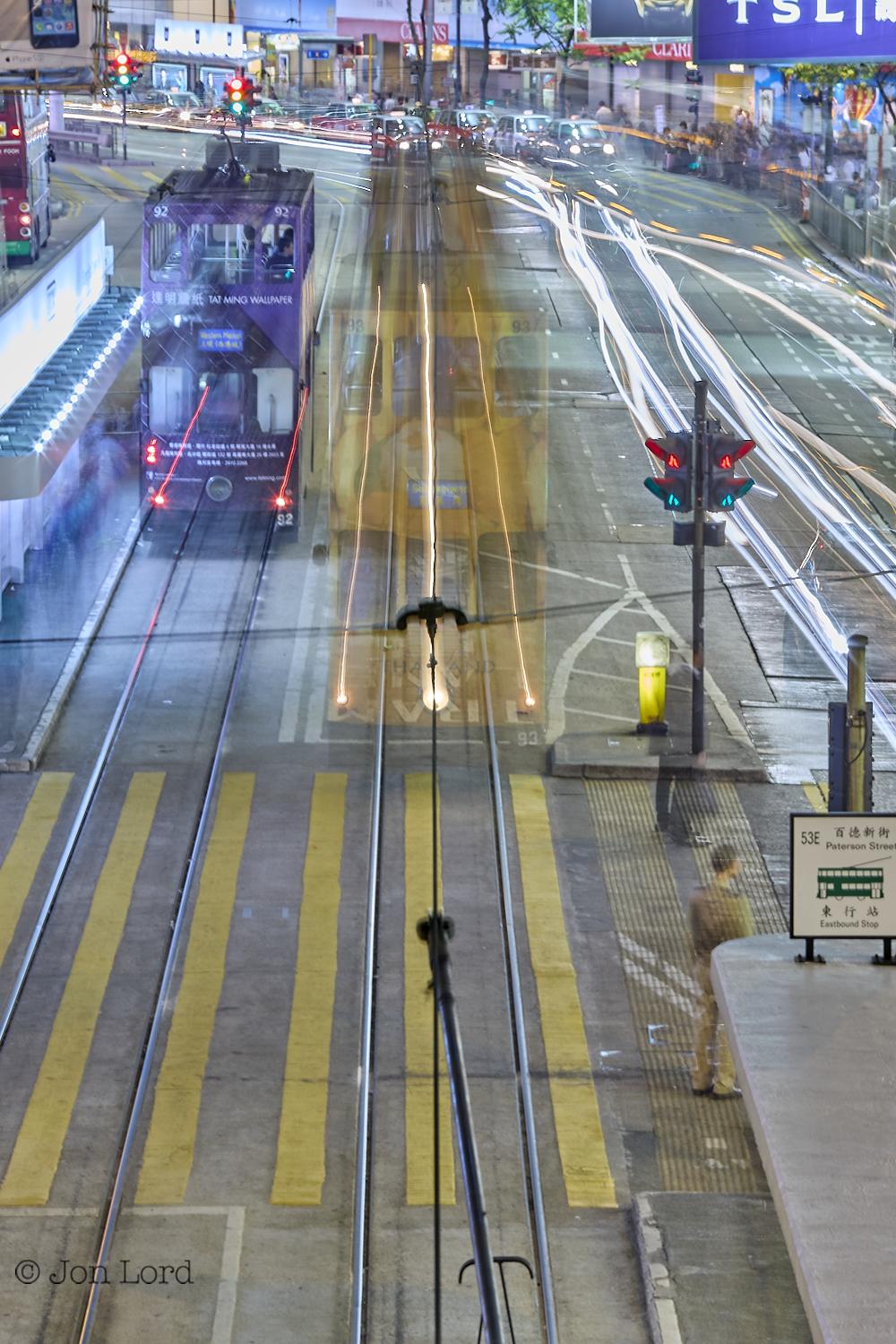 This is a colour, long exposure, night time street photo in portrait format of a city street scene photographed from above displaying motion blurred trams, cars and people. Causeway Bay, Hong Kong (2014).

The camera is positioned on an pedestrian bridge about five metres above the street and angled along a fifty metre, straight section of road before the road bends out view to the left. Under the camera and extending away into the distance is a pair of rail tracks embedded into the street. A few metres to the left is a further, parallel pair of tracks. In the centre of the image and partially blurred, is the front of a yellow double deck electric tram. The front of the tram is slightly transparent with parts of the street visible underneath. The dark, back of the tram extends away from the camera perhaps ten metres marking its passage along the street before it stopped. On the left side of tracks is a further tram, this one is purple and not as ghostly and transparent as the first. Th